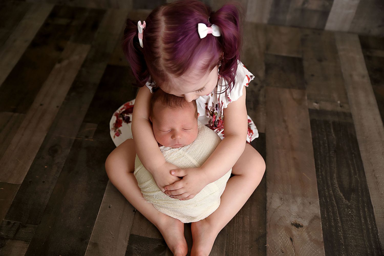 Young girl with purple pigtails sitting on wood floor gently holding a sleeping newborn baby sibling.
