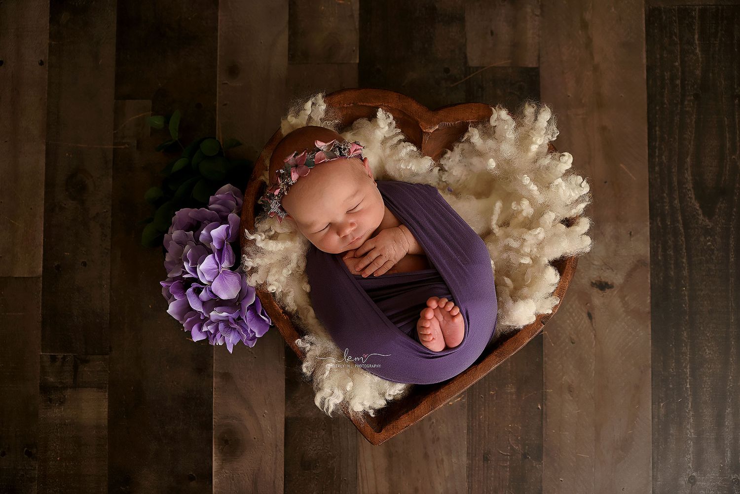 Sleeping newborn baby wrapped in purple swaddle with matching headband posed in a heart-shaped wooden bowl.