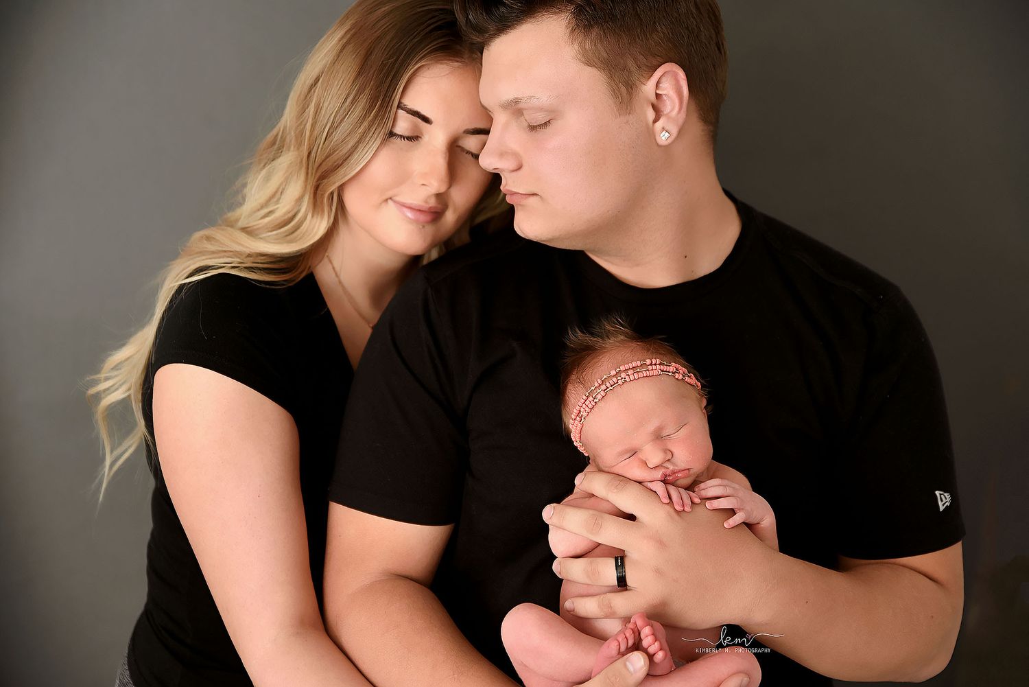 New parents dressed in black tenderly holding their sleeping newborn baby together.