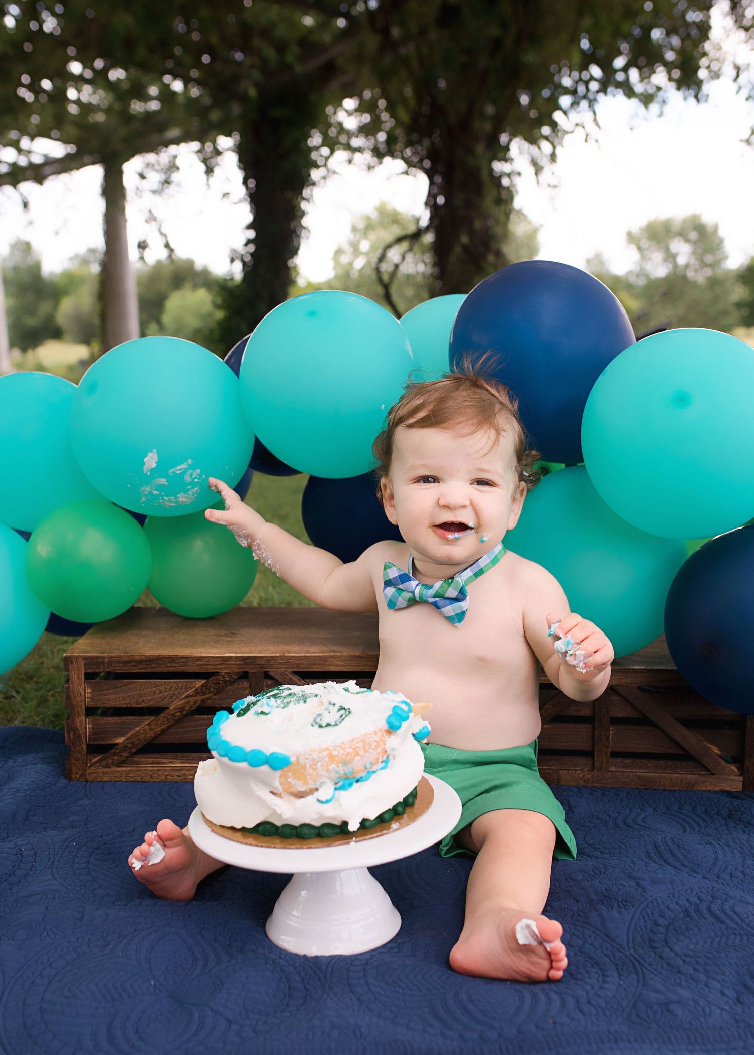 First birthday celebration setup with teal and navy balloons, wooden crate, and white frosted cake on blue blanket.