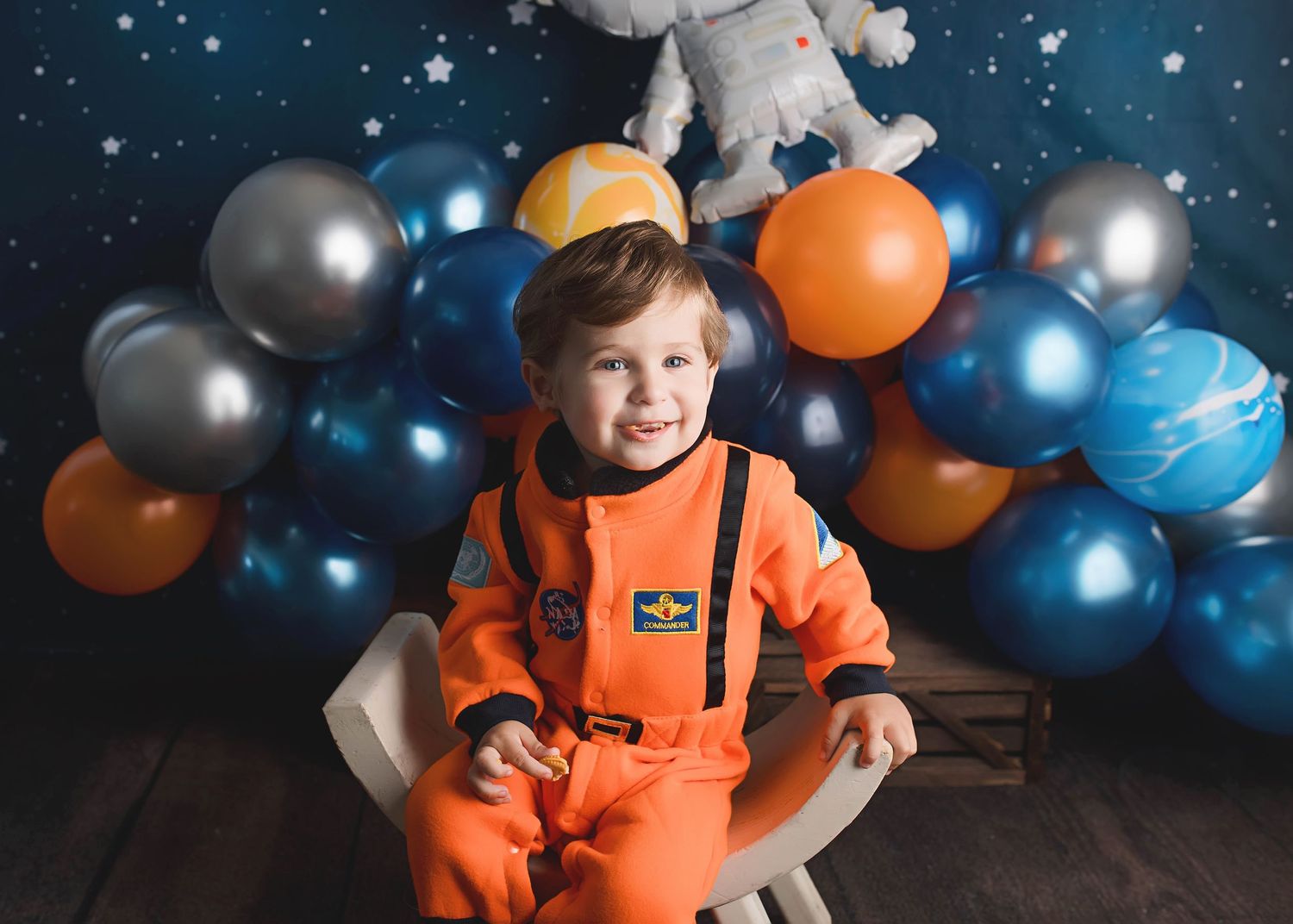Space themed first birthday photo with orange astronaut costume and blue balloon decorations.