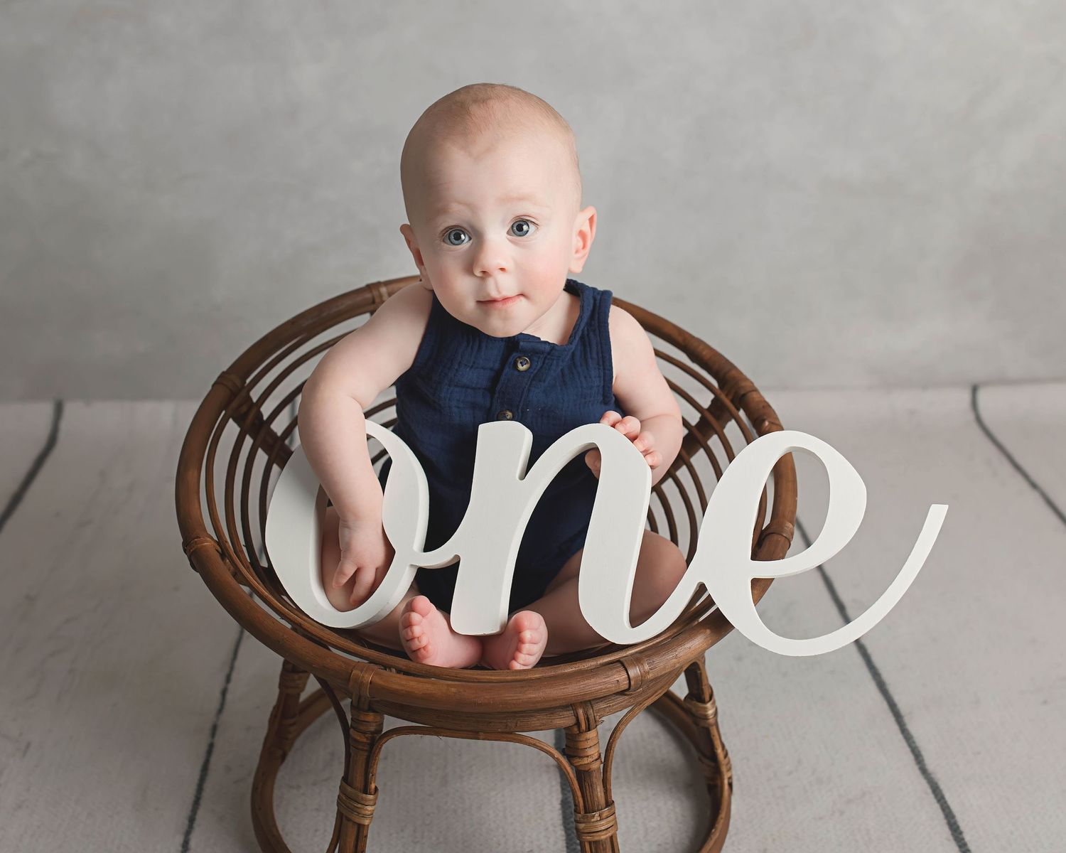 Minimalist first birthday photo with wooden chair and 'one' text prop against grey background.