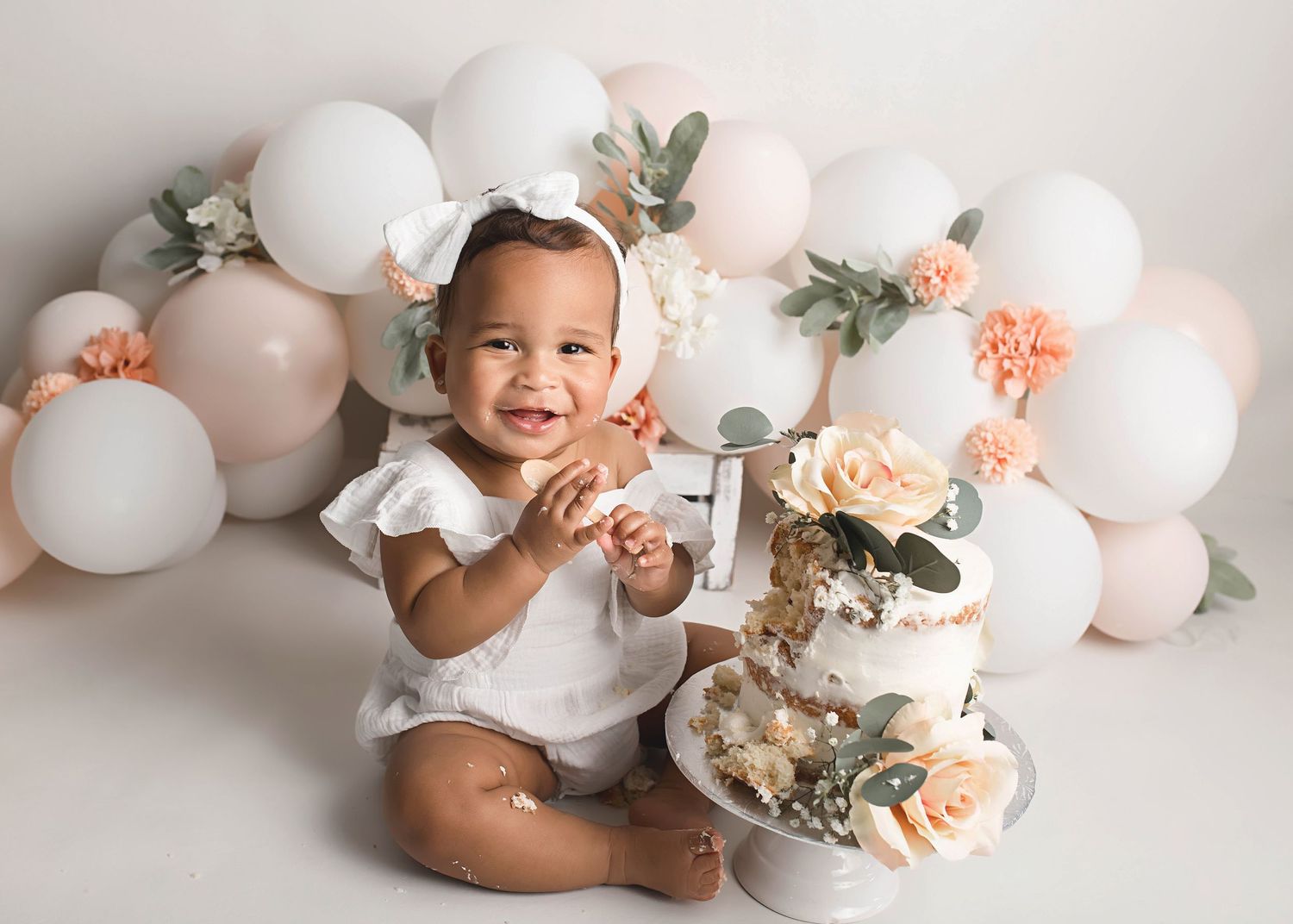 Smiling baby next to first birthday cake with white and peach balloon garland.