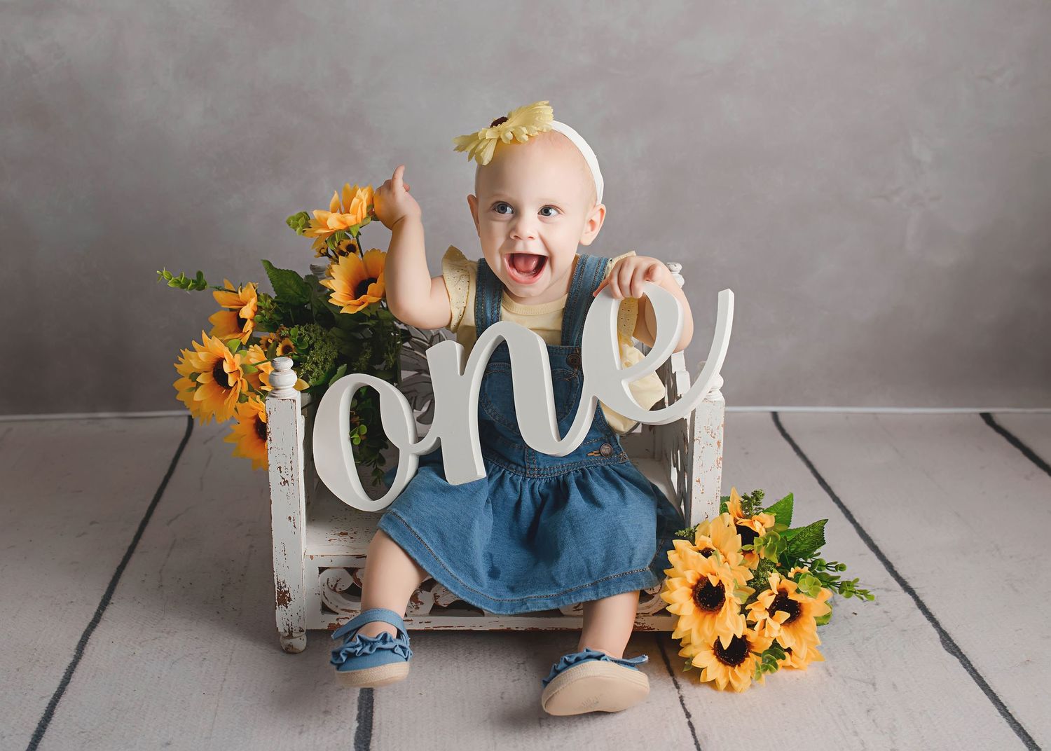 Toddler in blue pants sits with 'one' text display and sunflowers for first birthday photo.