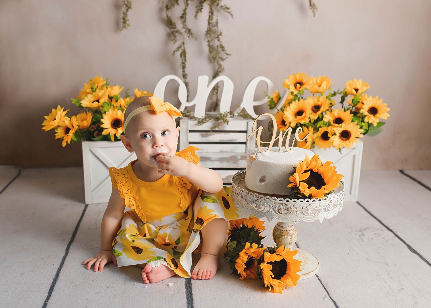 Baby in mustard yellow outfit poses with sunflowers and 'one' text display.