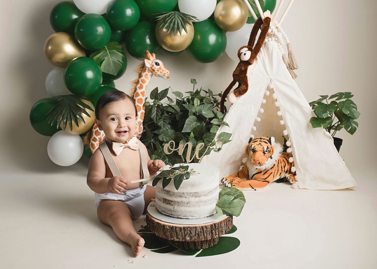 Safari themed first birthday setup with green and gold balloons, teepee, and cake.