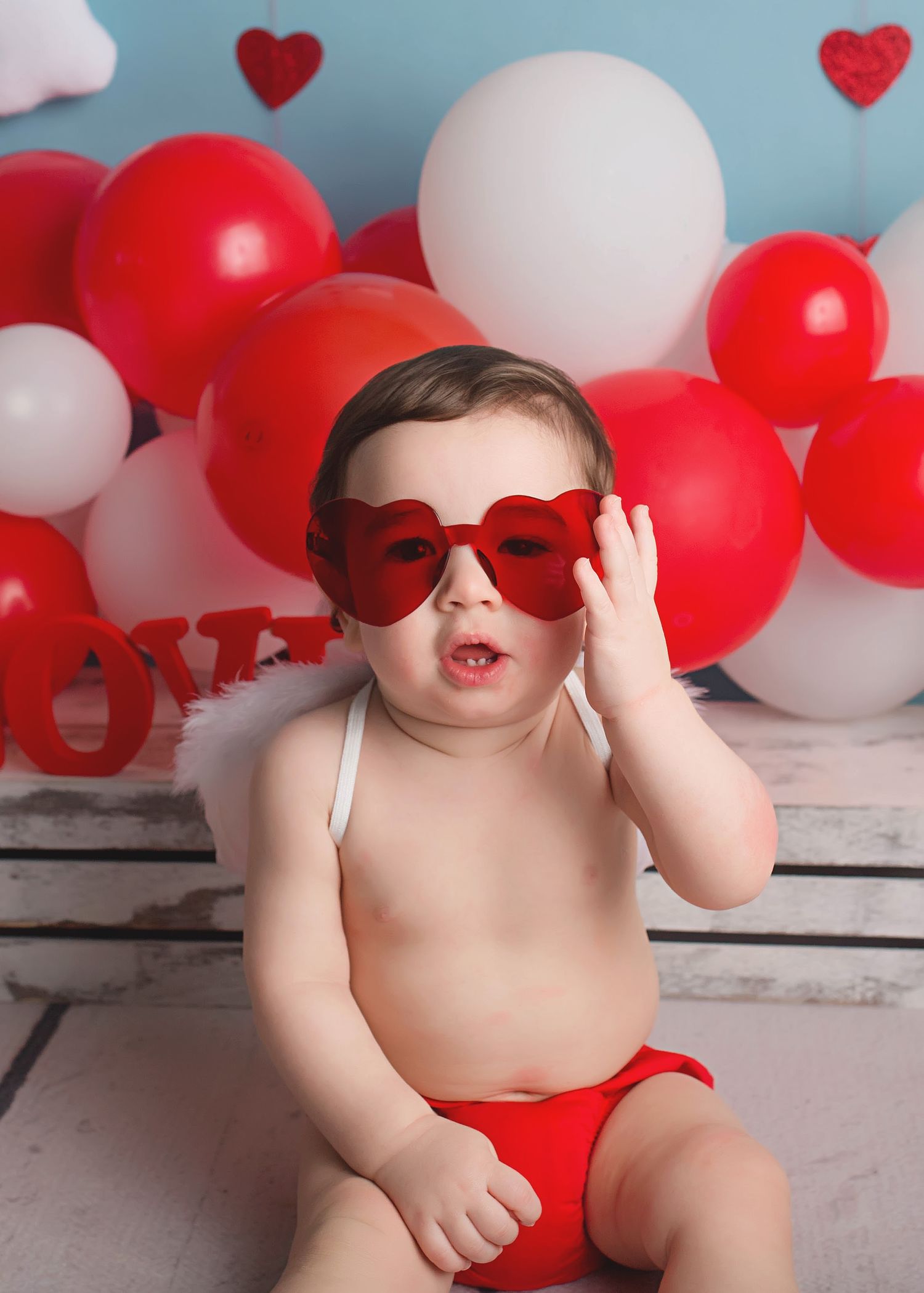Baby in red diaper wearing heart sunglasses against Valentine backdrop.