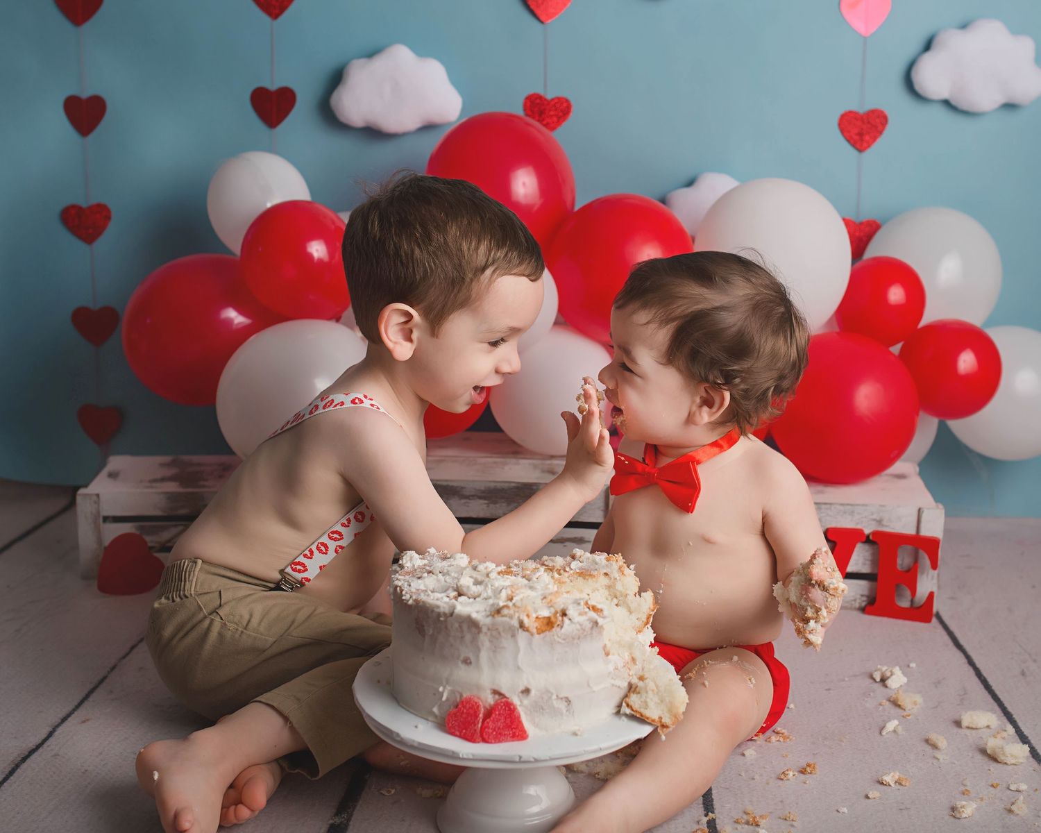 Two children sharing a white birthday cake with heart balloon decorations.