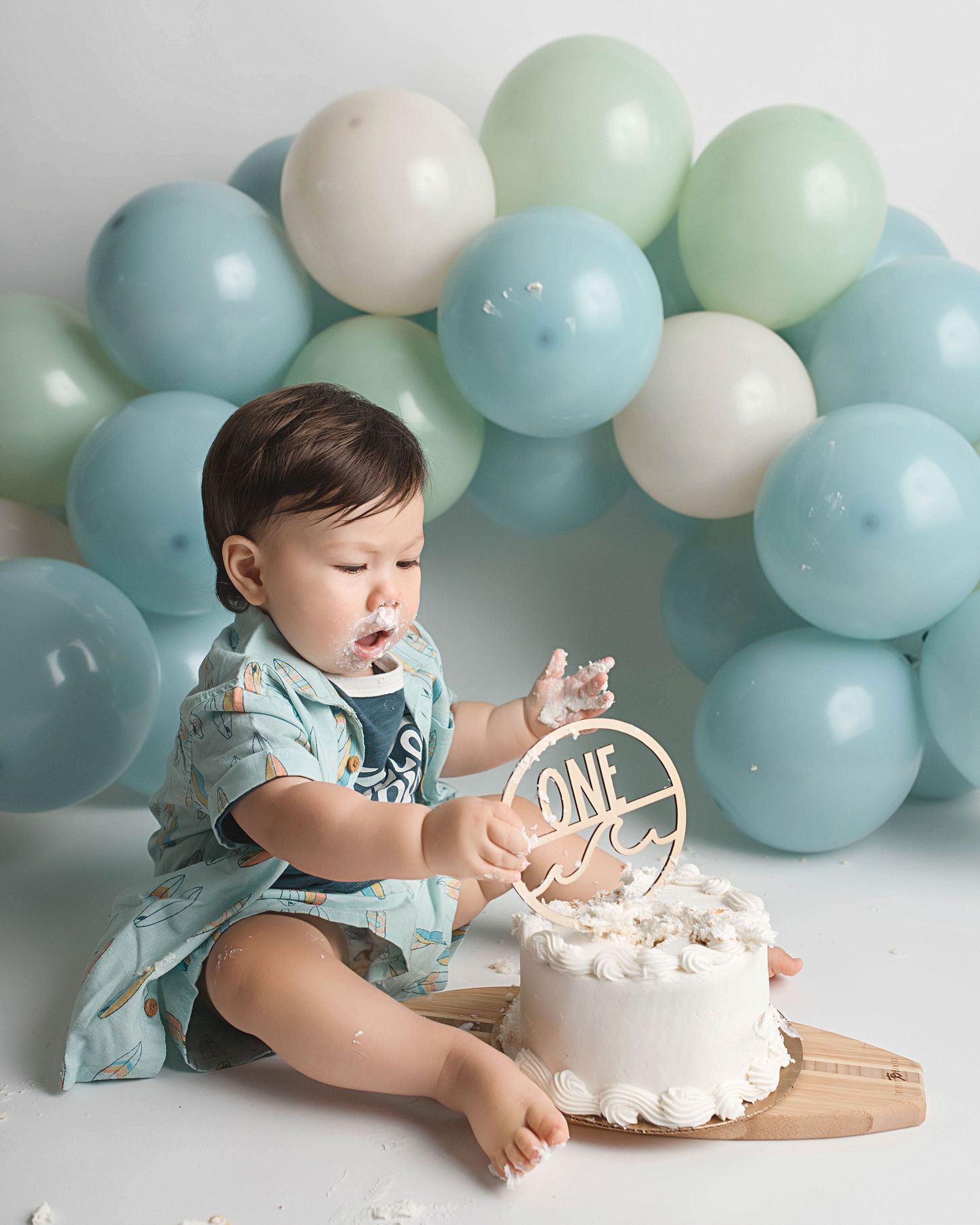 Baby reaching for a white birthday cake with 'ONE' topper surrounded by blue balloons.