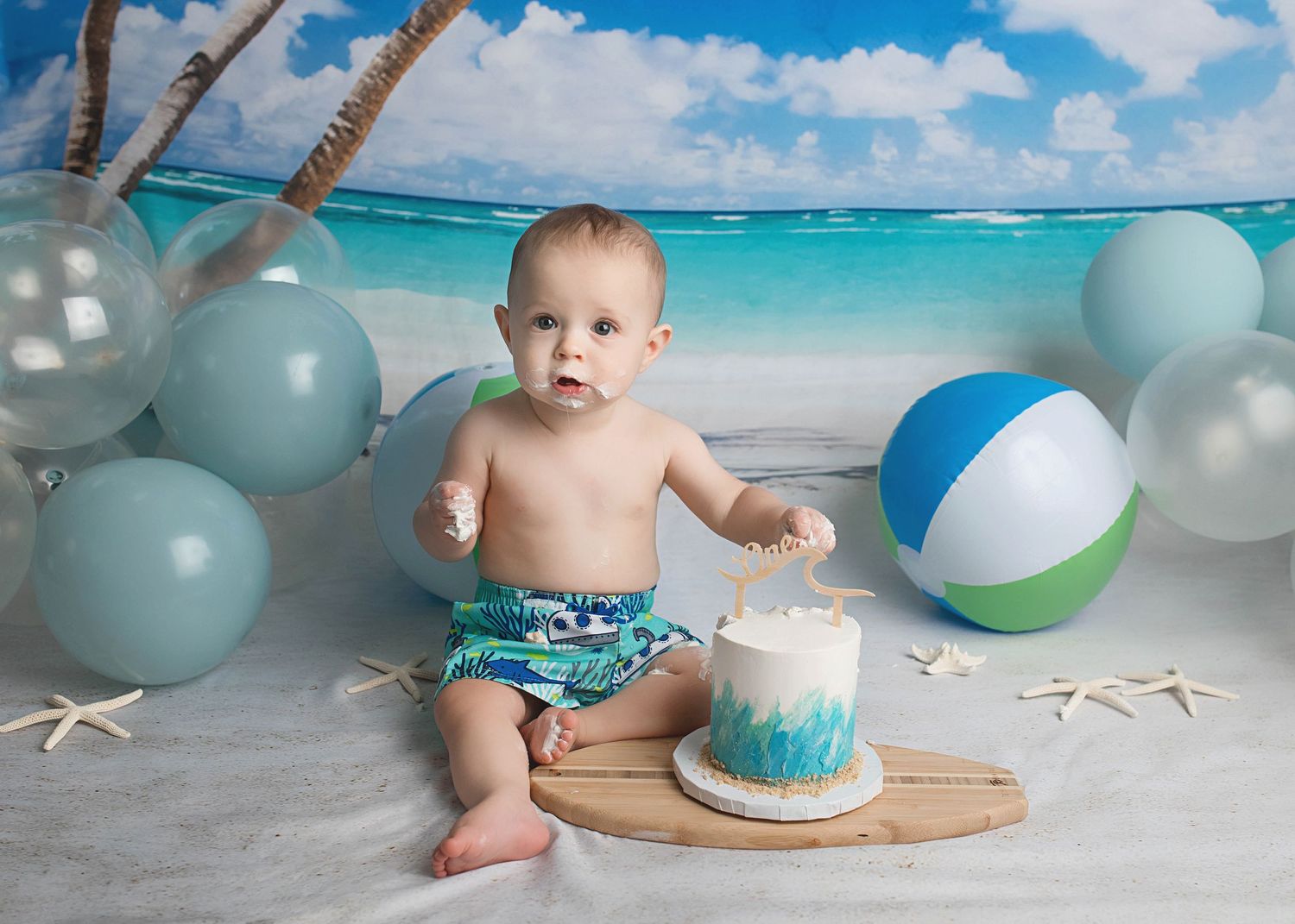 Beach themed cake smash photo with blue balloons and tropical backdrop.
