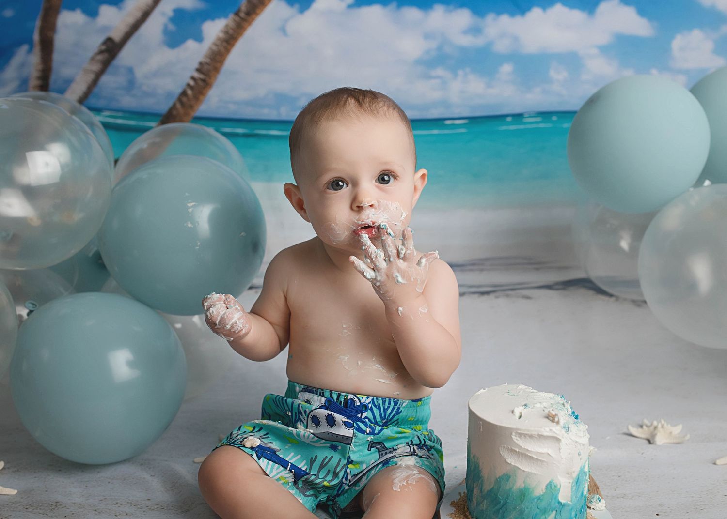 Baby enjoying cake during beach themed first birthday photoshoot with blue balloons.