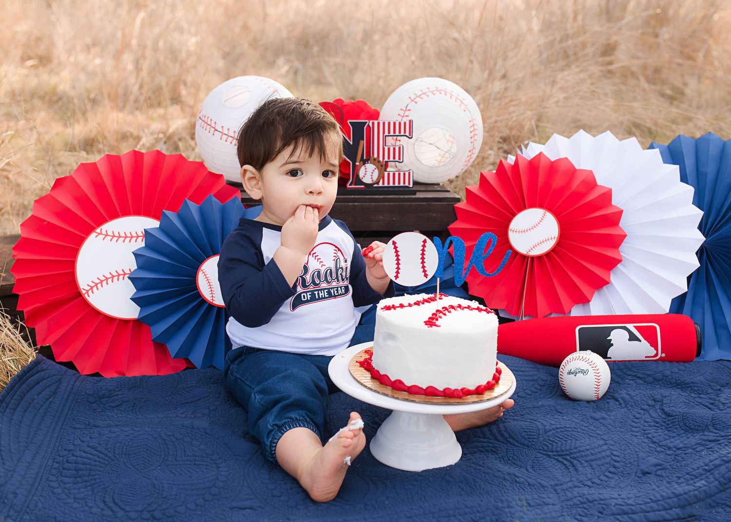 Baseball themed first birthday setup with red and blue paper fans and white cake on navy blanket.