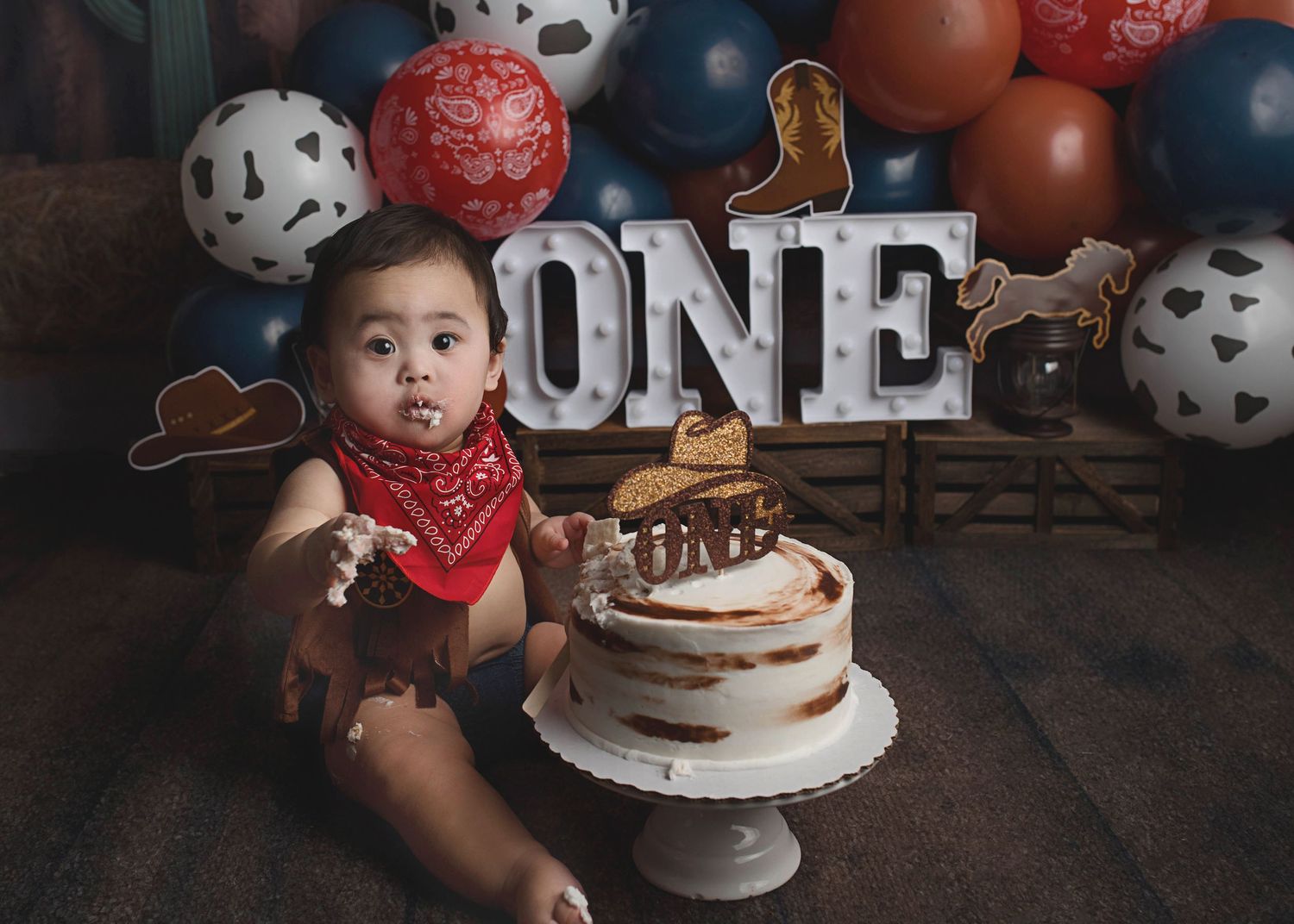Baby with red bandana sitting near first birthday cake with western themed decorations.