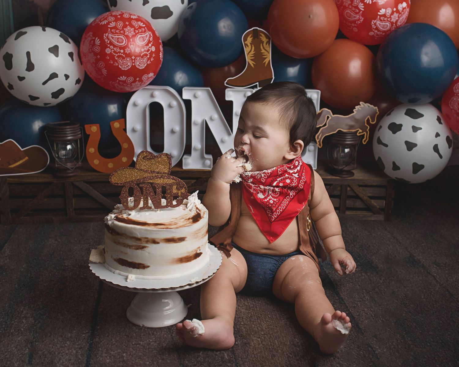 Baby in red bandana reaching for birthday cake during western themed photo session.