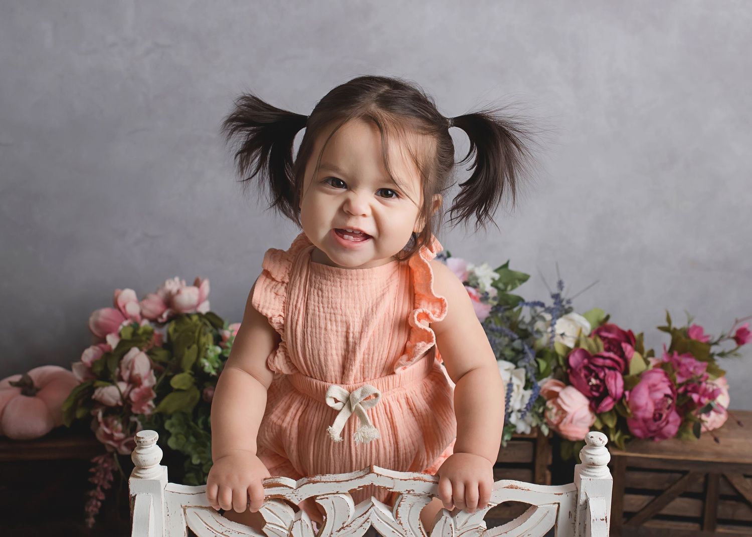 Portrait of toddler in peach dress sitting in white vintage chair with floral backdrop.