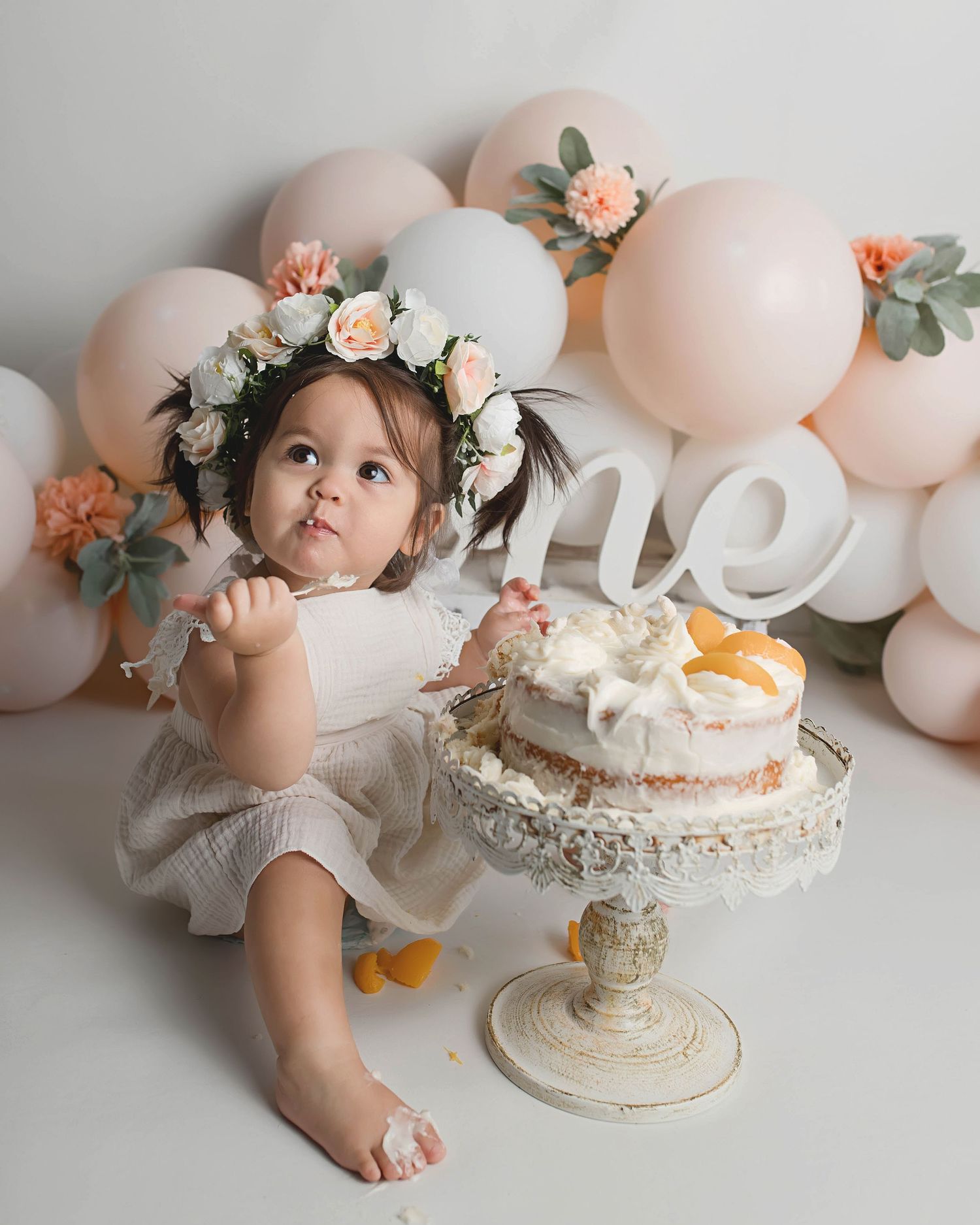 First birthday cake smash scene with floral crown, pink balloons and elegant white cake stand.