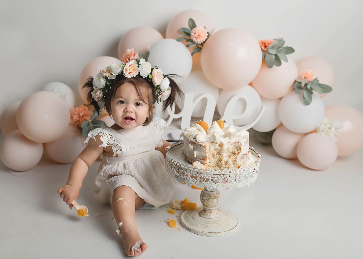 First birthday celebration setup with pink balloons, floral crown, and white cake on ornate stand.