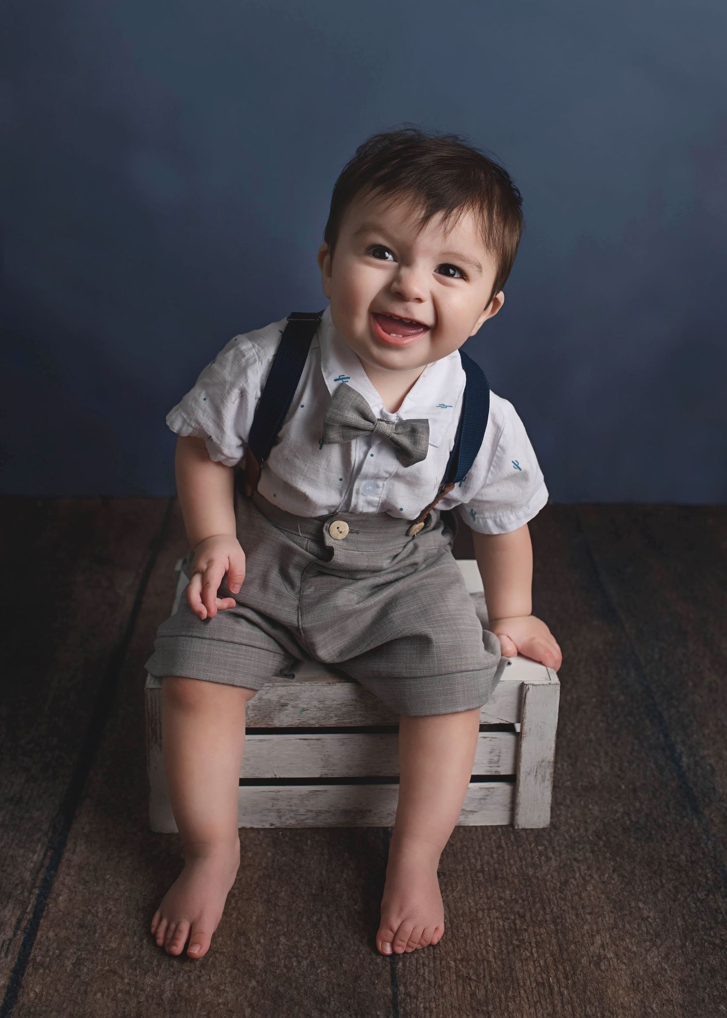 Toddler in vintage-style outfit with suspenders and bowtie sits on wooden crate.