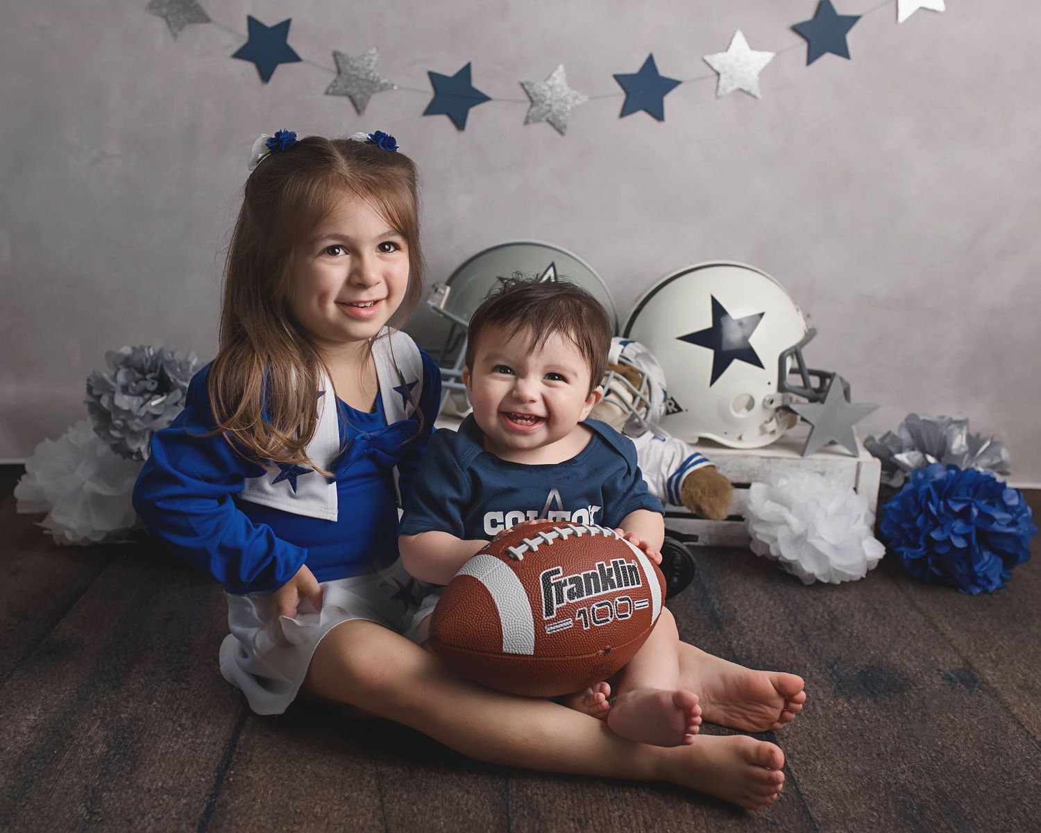 Two children sit on wooden floor with football and star decorations against gray backdrop.