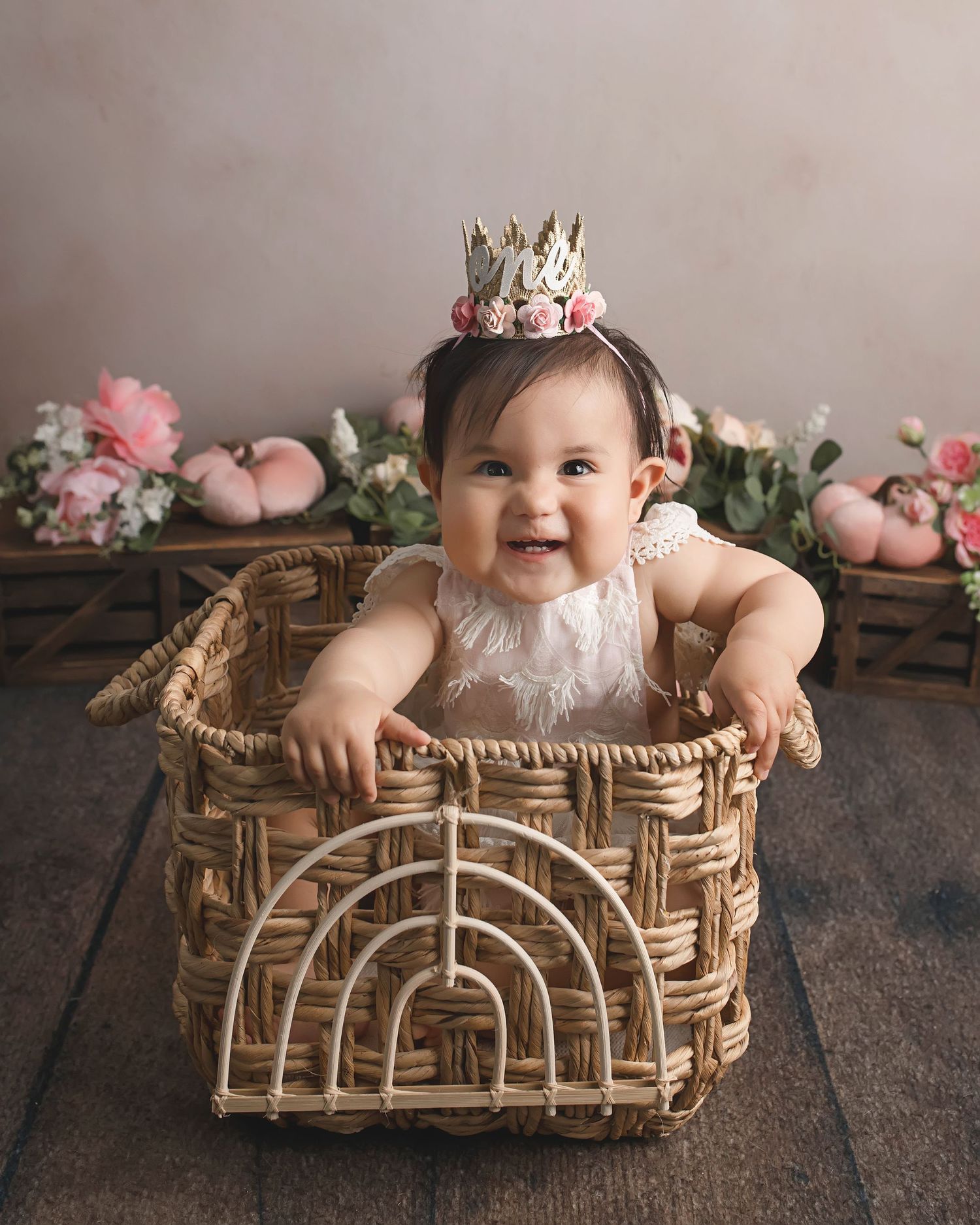 Baby in white dress and crown sitting in decorative wicker basket with floral accents.