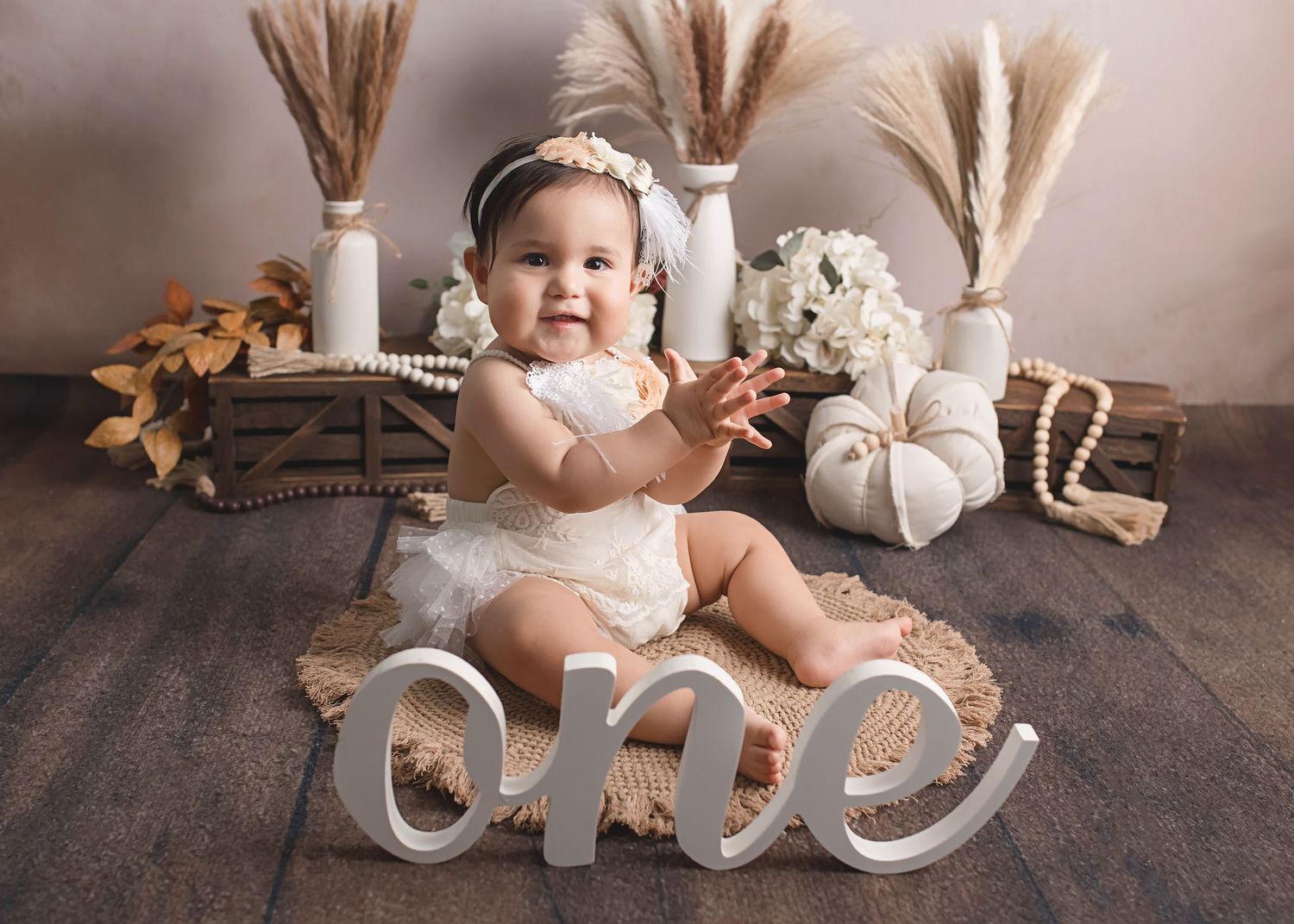 Baby in white dress posing with decorative 'one' sign and neutral boho decor.