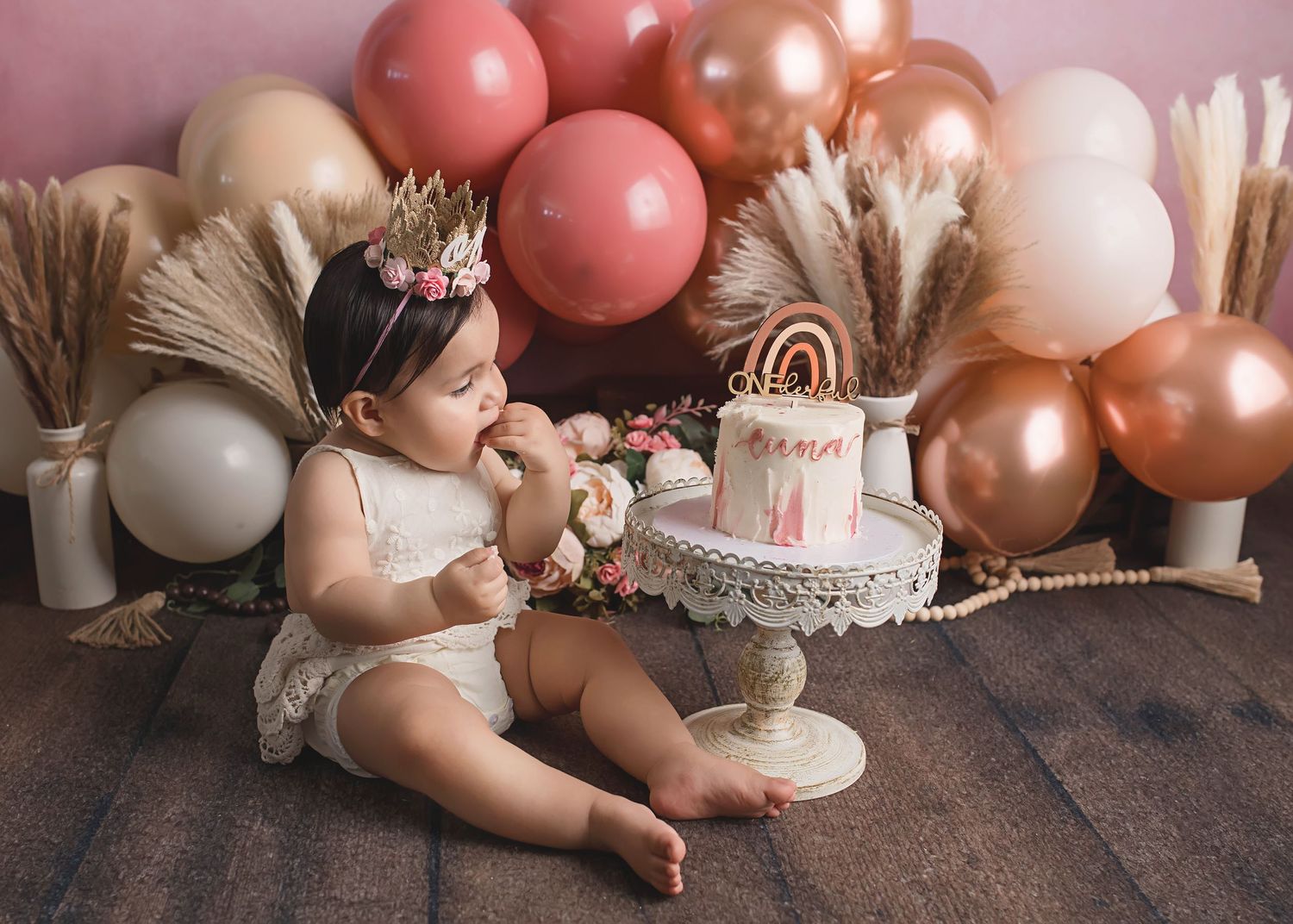 First birthday cake smash scene with pink balloons, pampas grass, and tiered cake.