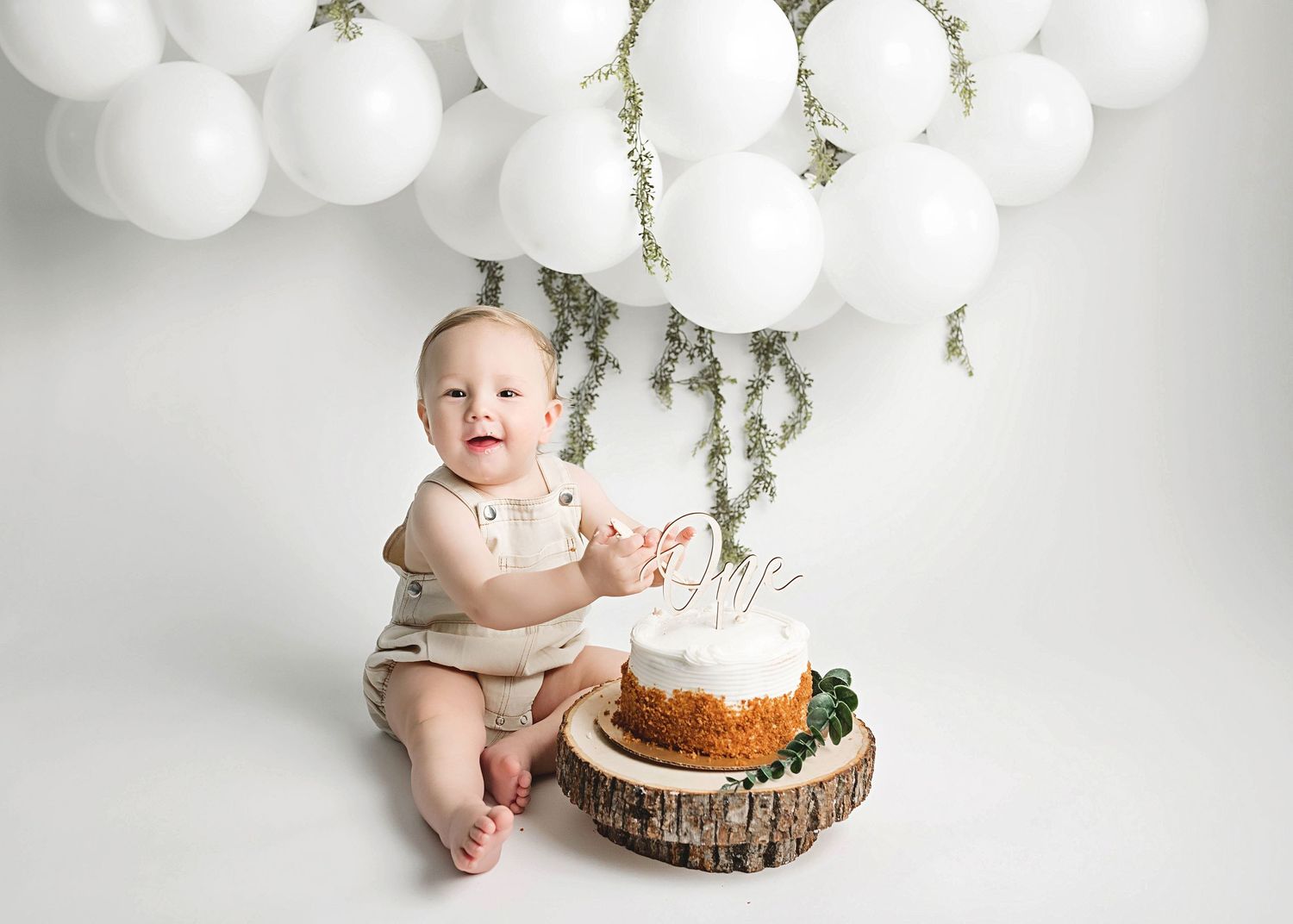 Baby wearing light colored outfit sitting next to first birthday cake with white balloon decorations.