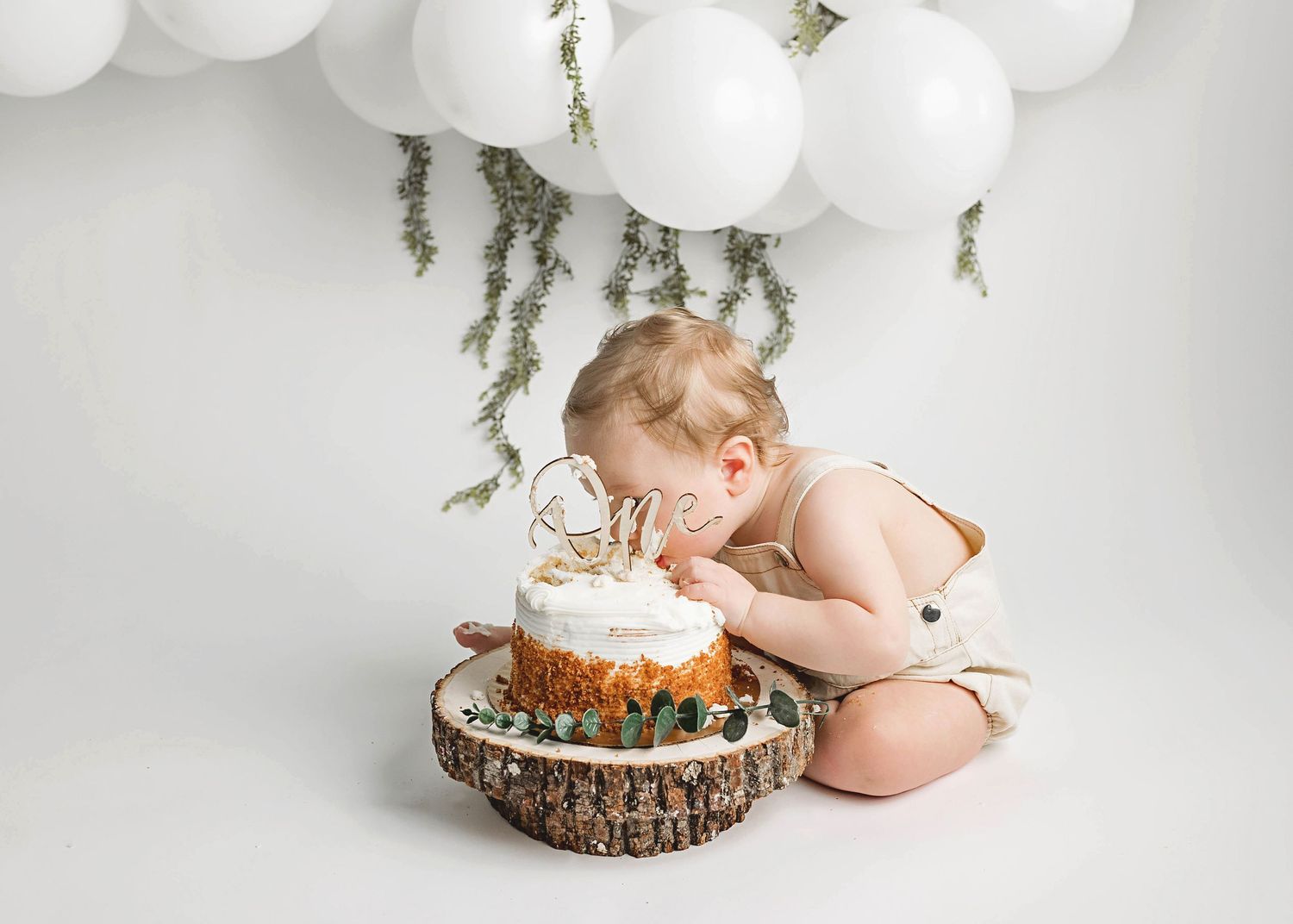 Baby in white outfit exploring birthday cake on rustic wood slice with white balloon and greenery backdrop.