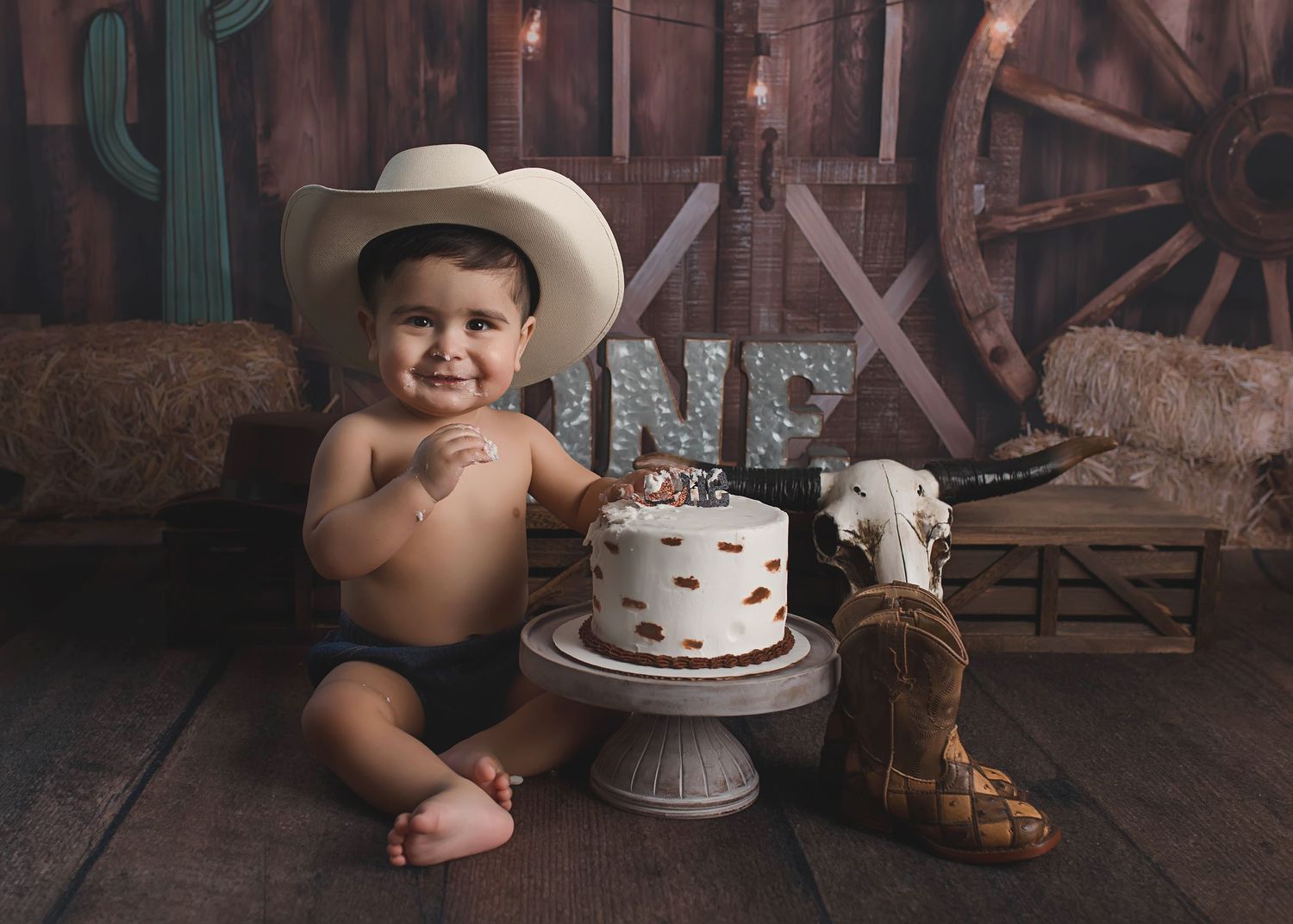 A smiling baby in western attire with birthday cake and rustic backdrop.