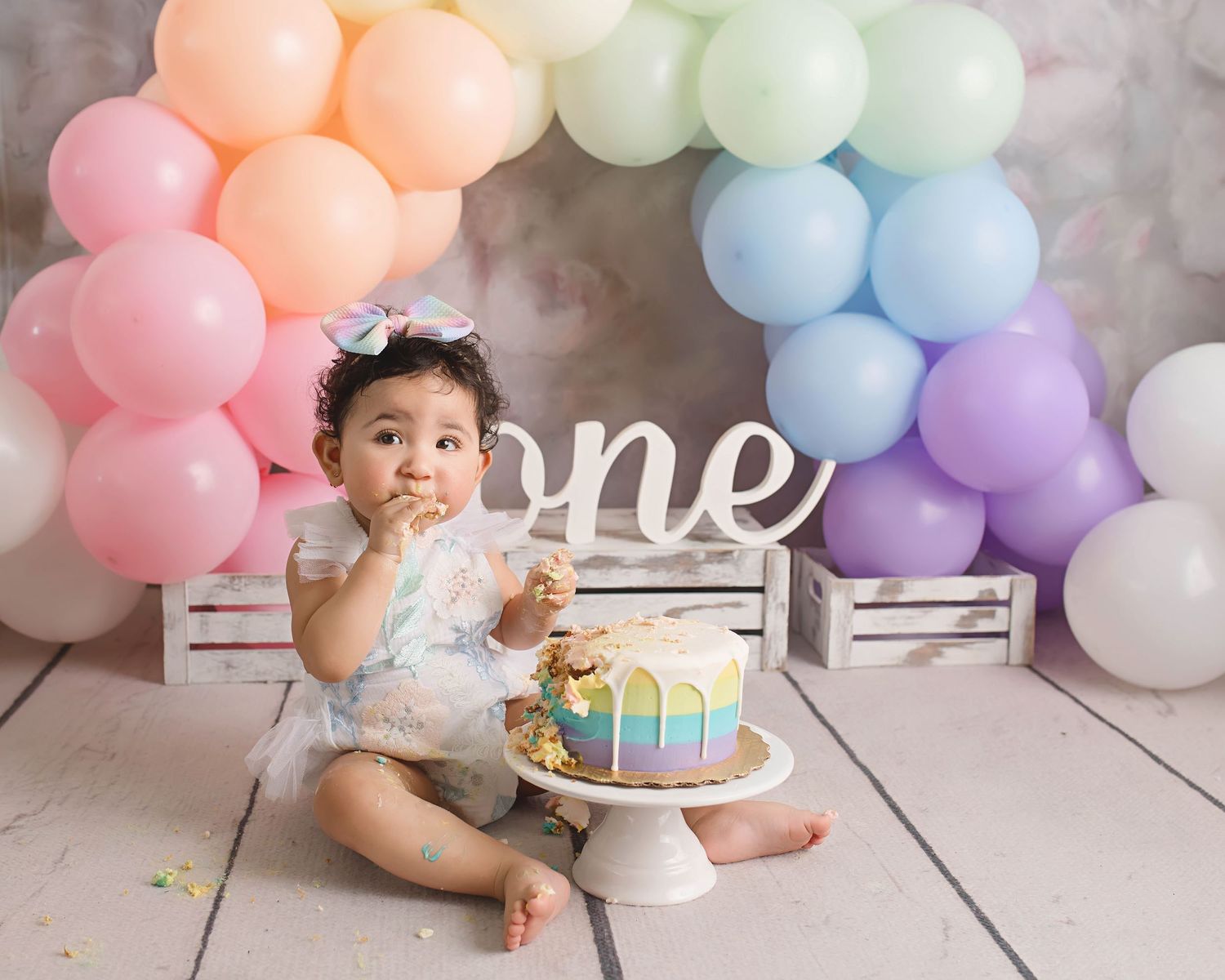 Pastel rainbow balloon arch backdrop with white 'one' sign for first birthday photoshoot.