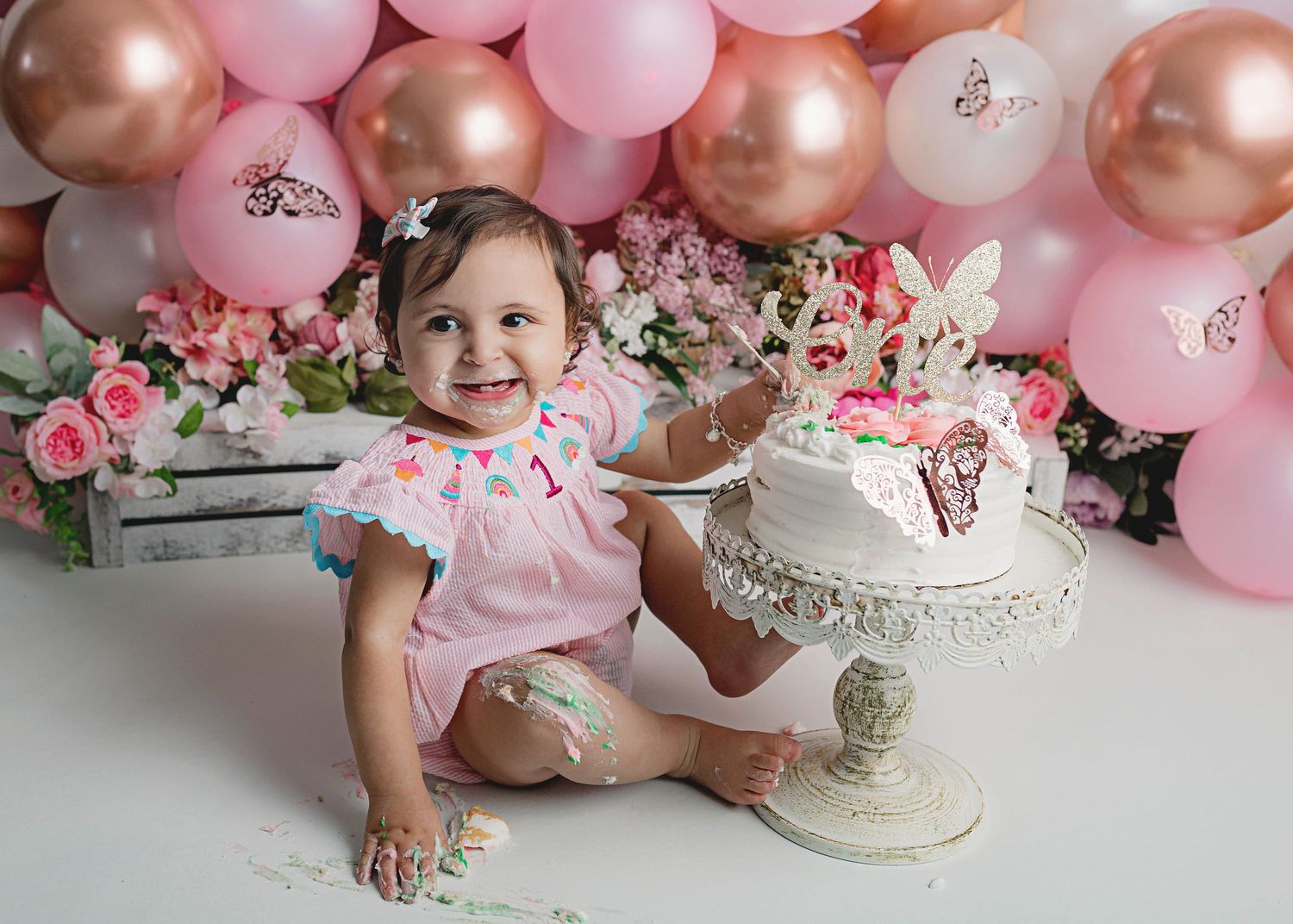 Smiling toddler in pink outfit next to decorated cake stand with rose gold and pink balloon backdrop.