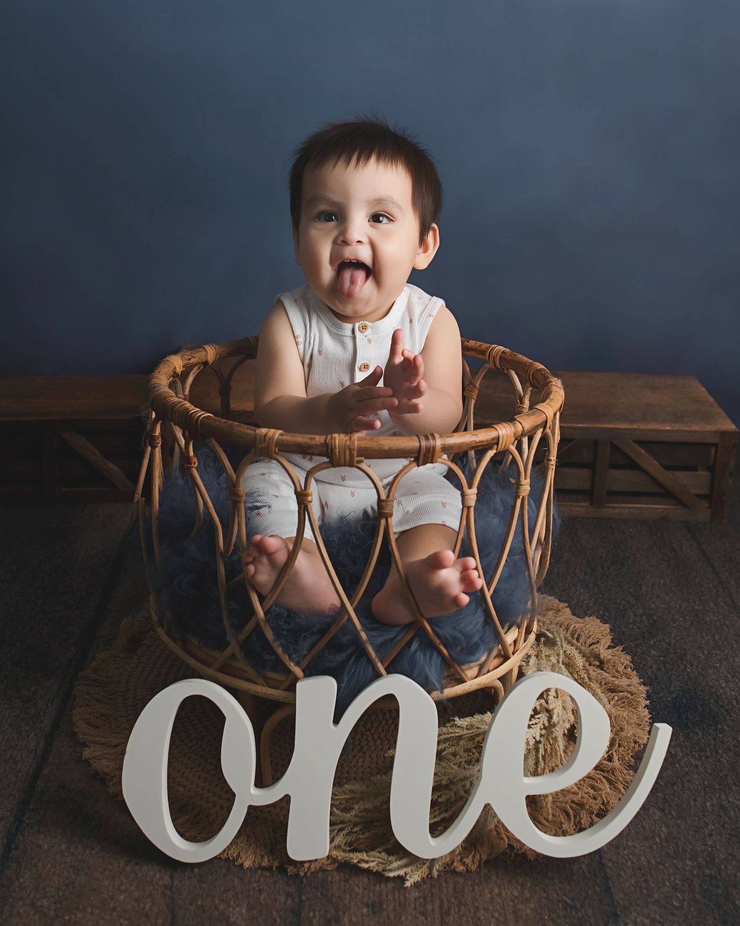 Baby sitting in wooden basket with 'one' text sign for first birthday milestone photo.