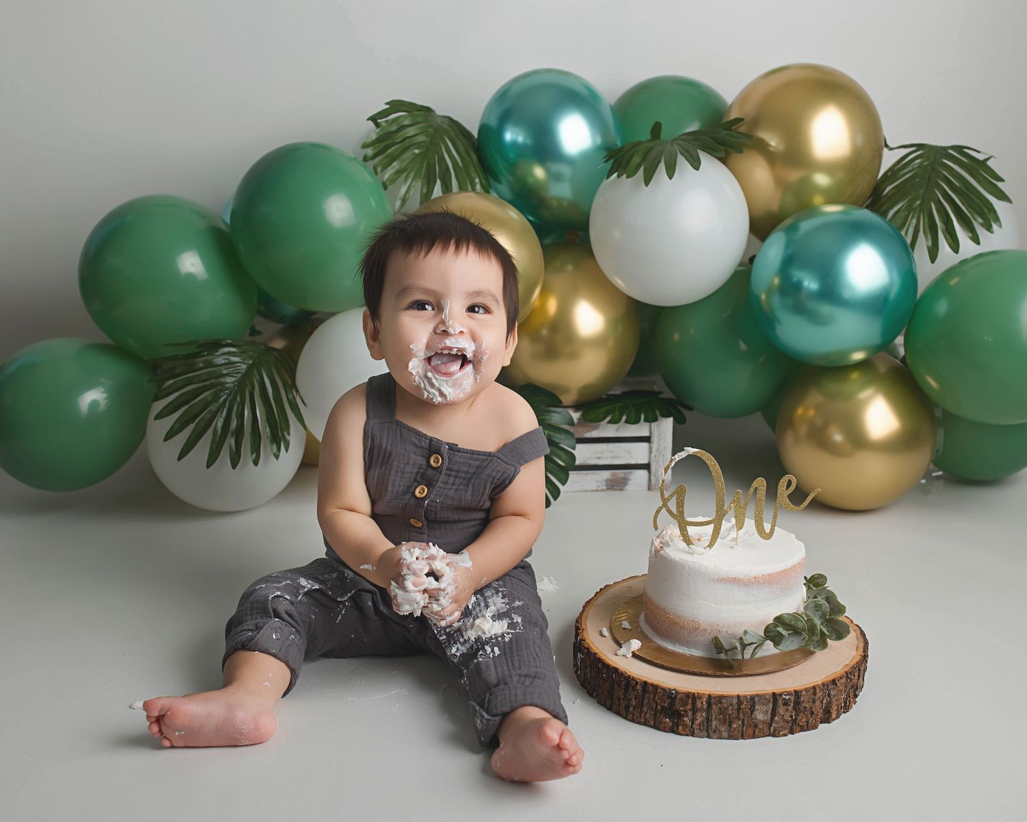 One year old enjoying cake smash session with green and gold balloon backdrop.
