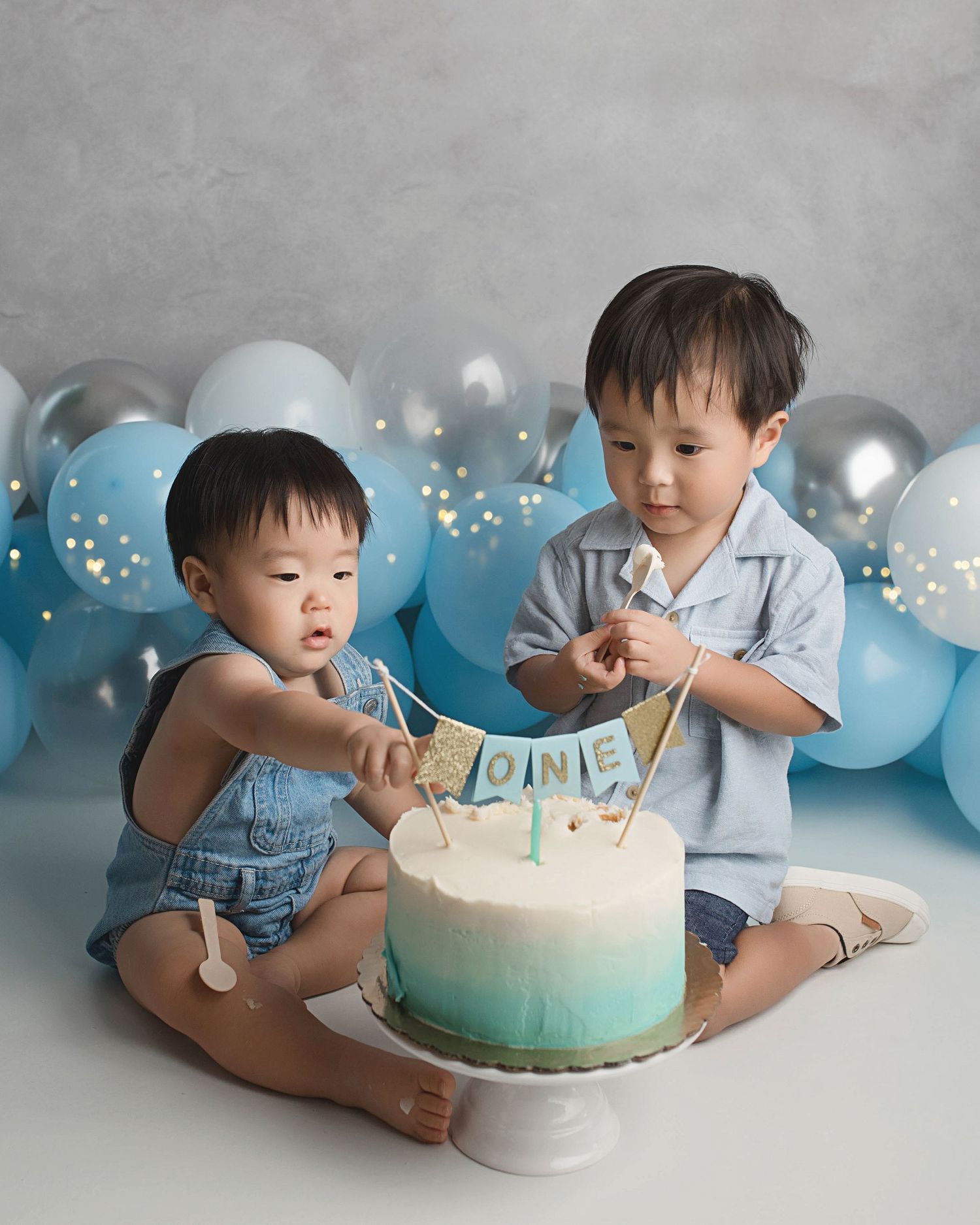 Two children sitting with ombre blue birthday cake surrounded by balloons.