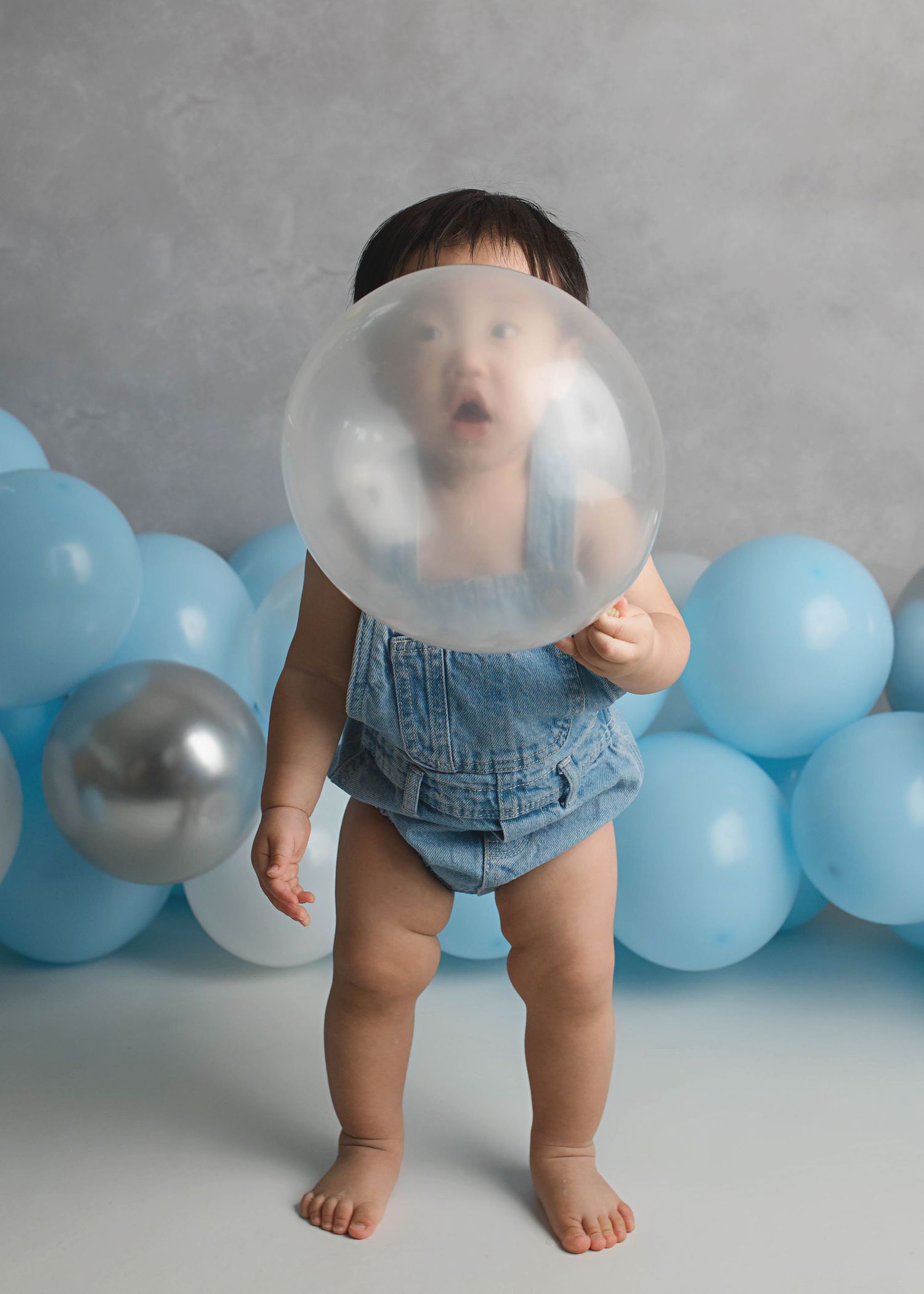 Baby in denim romper playing with bubble against blue balloon backdrop.