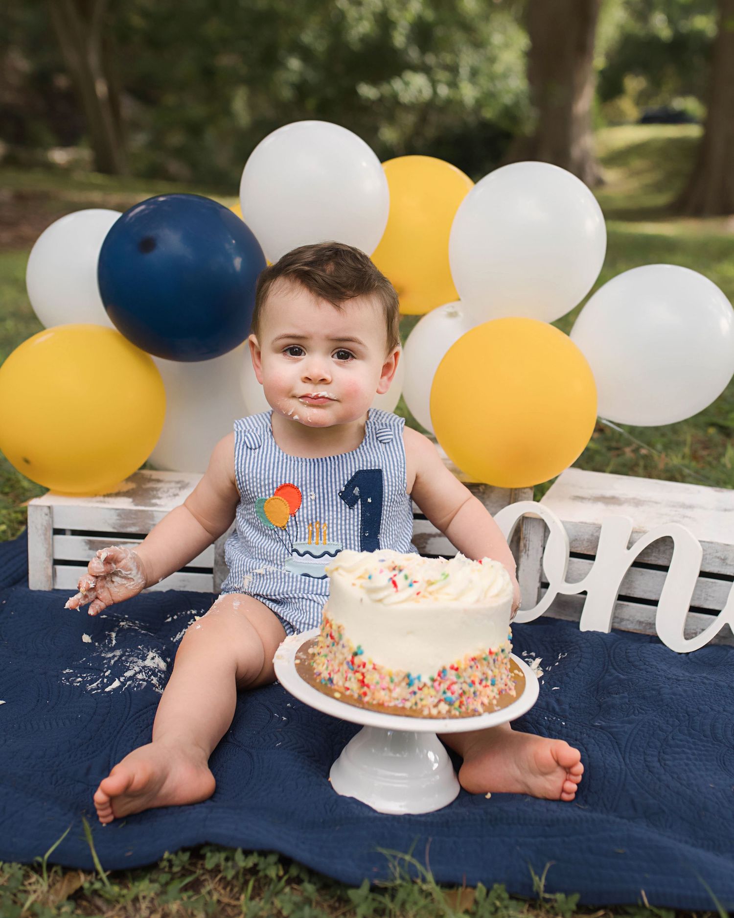 First birthday celebration setup with blue and yellow balloons surrounding a smash cake on navy blanket.