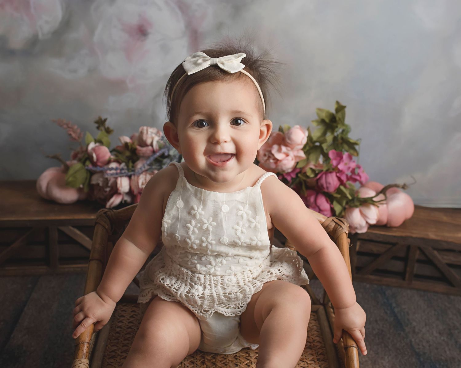 Baby in white lace romper sits in wooden chair surrounded by pink flowers and grey backdrop.