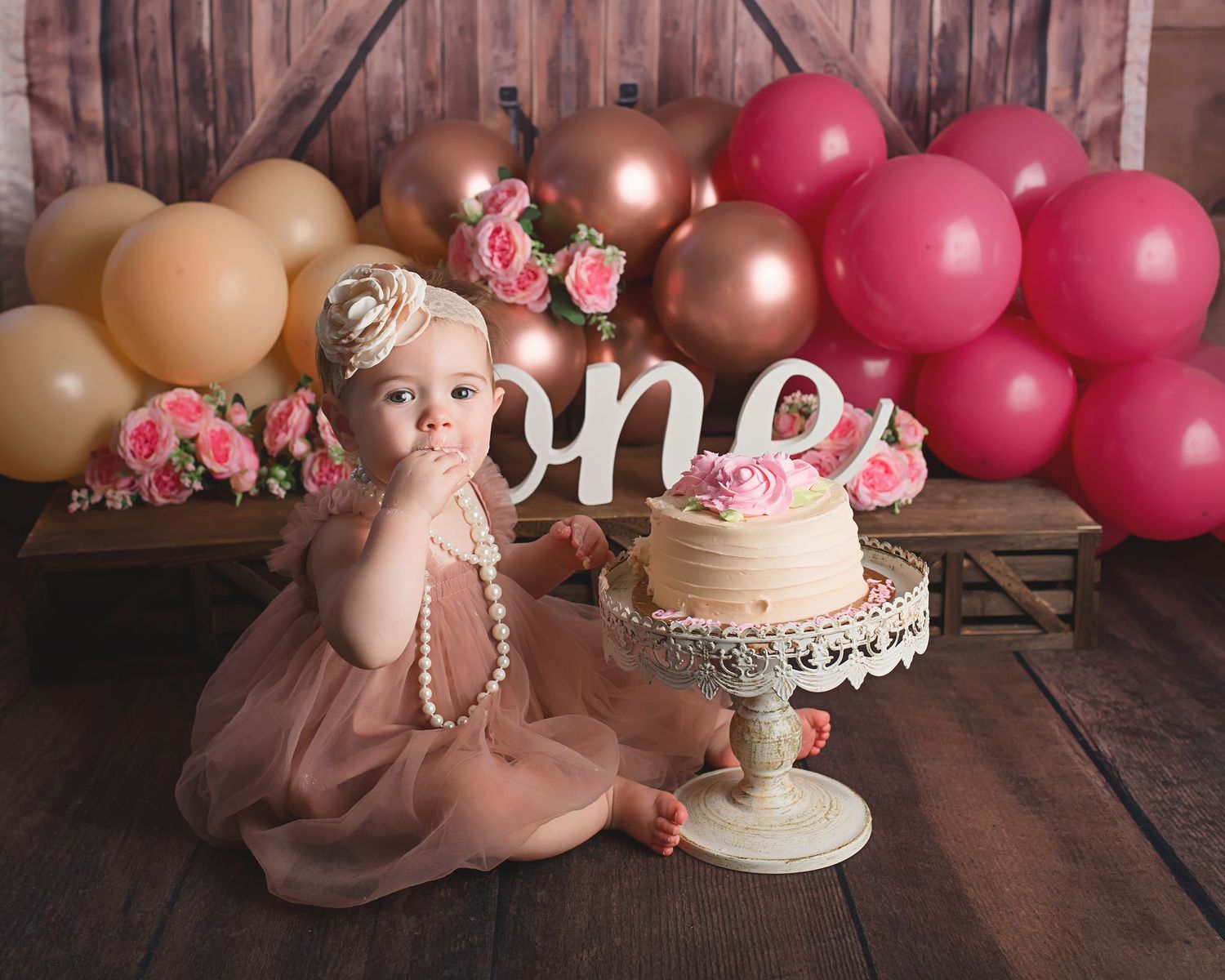 First birthday setup with ombre balloon arch, roses, and cake against rustic wooden backdrop.