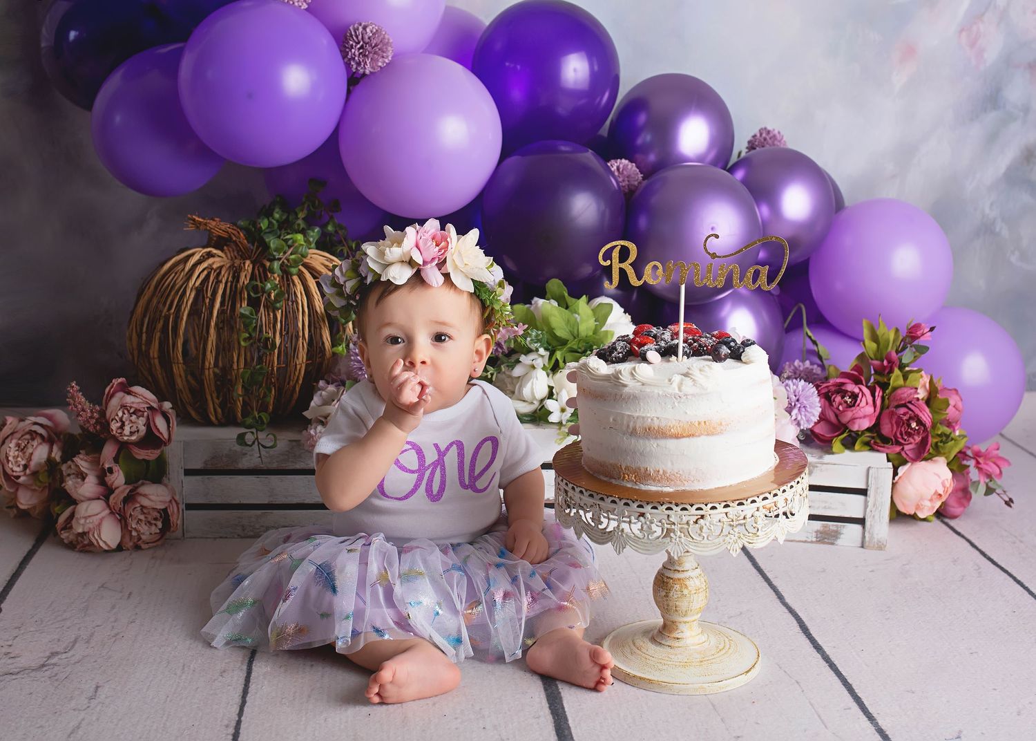 Birthday setup with purple balloon arch, floral decorations and cake on white pedestal against light backdrop.