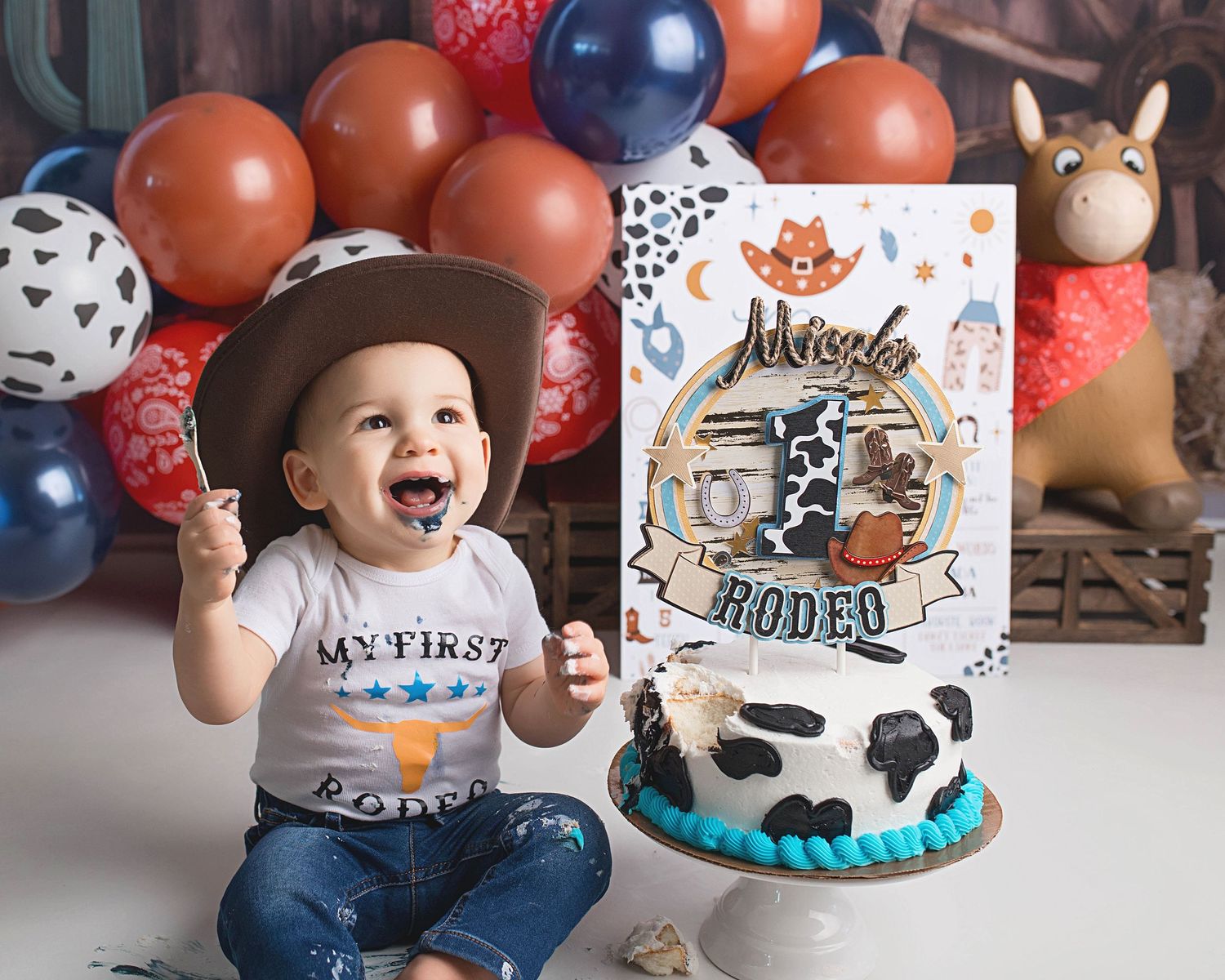 First rodeo birthday celebration with themed cake, balloons and excited baby in cowboy hat.