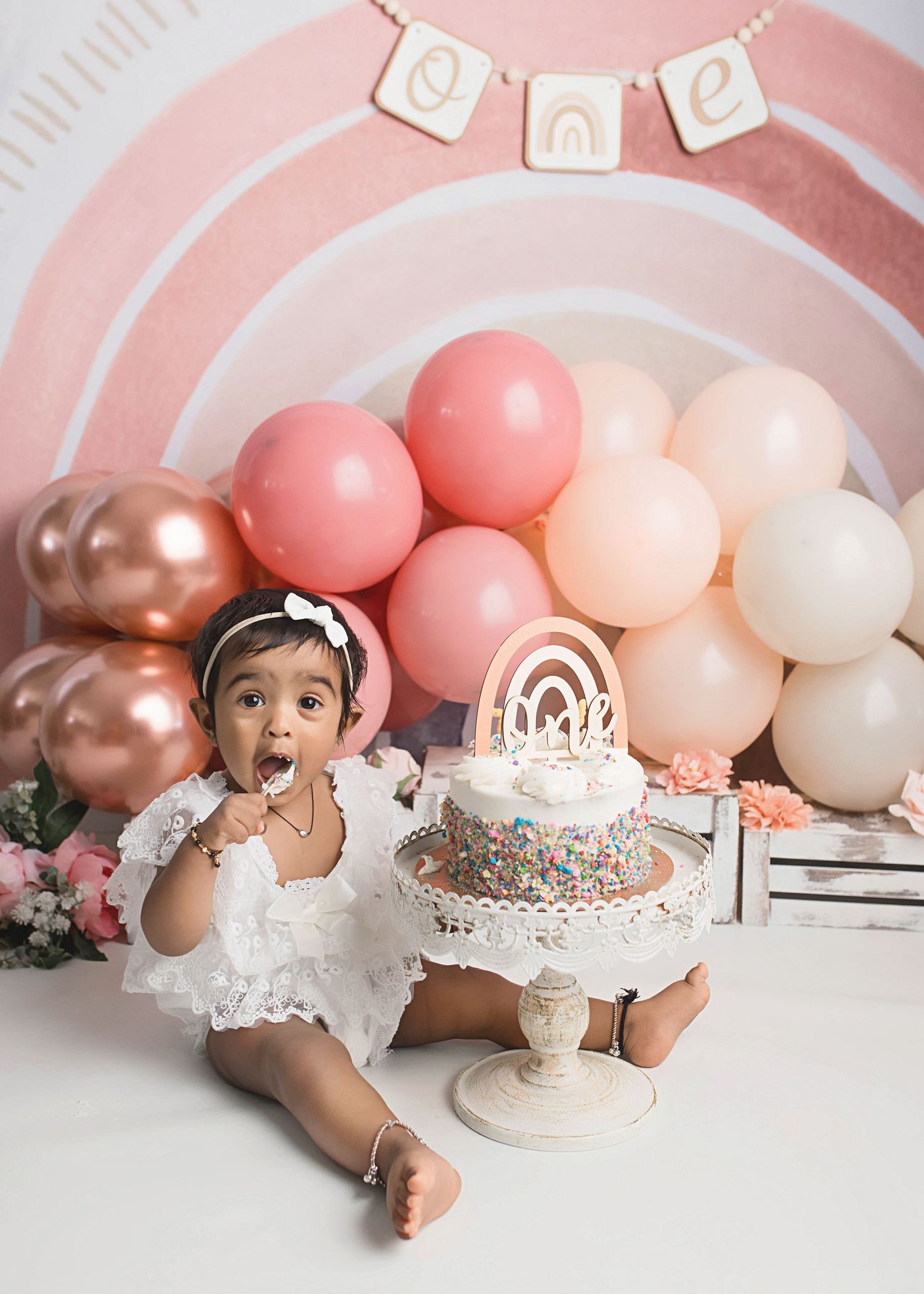First birthday celebration with pink balloon arch and white cake on pedestal.