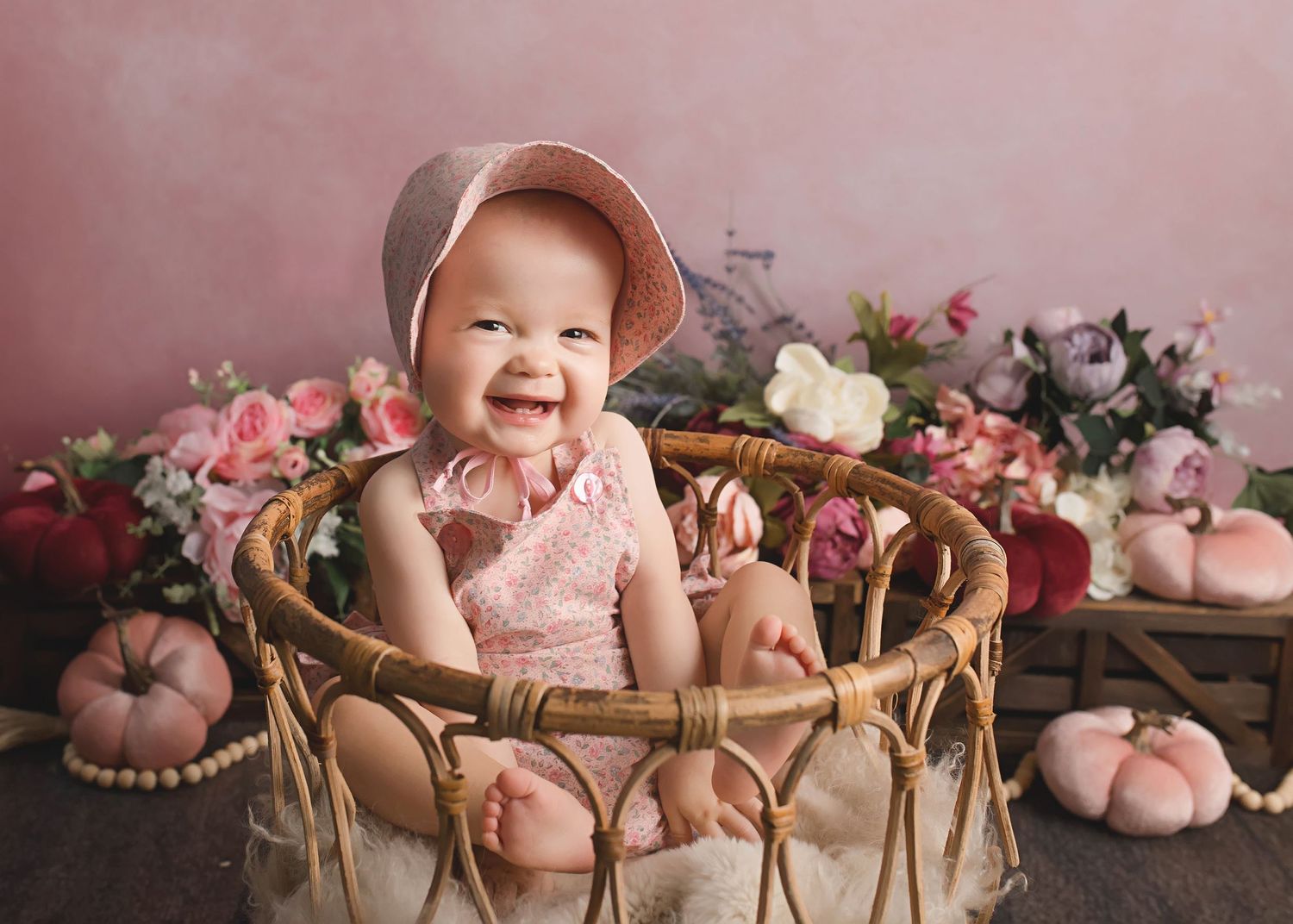 Baby in pink dress sitting in woven basket with floral backdrop.