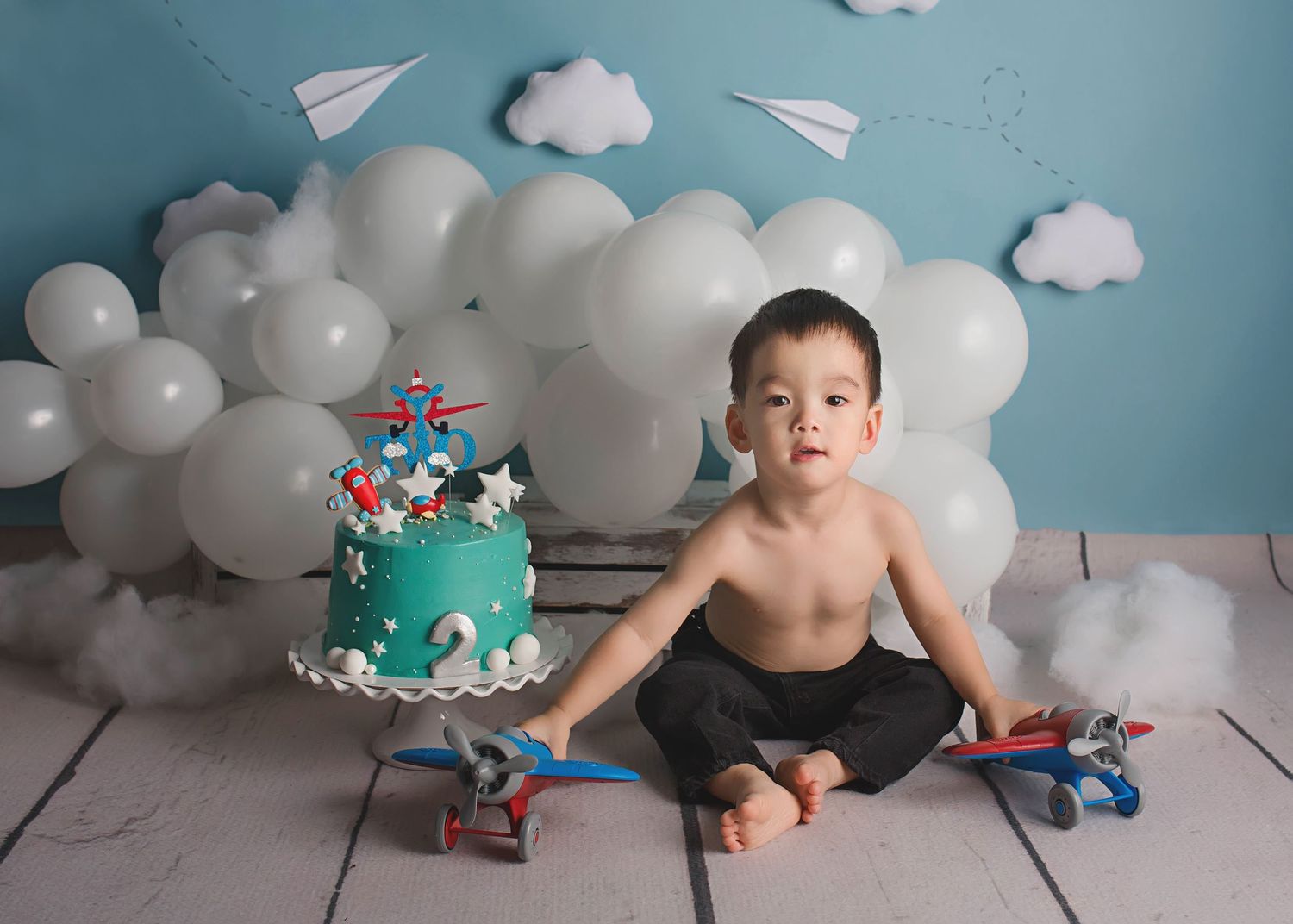 Aviation themed cake setup with white balloon clouds and paper airplanes on blue background.