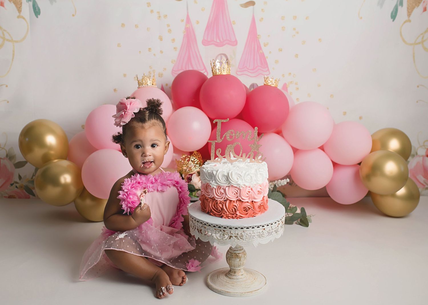 First birthday portrait with pink balloon arch, gold accents, and ombre rose cake.