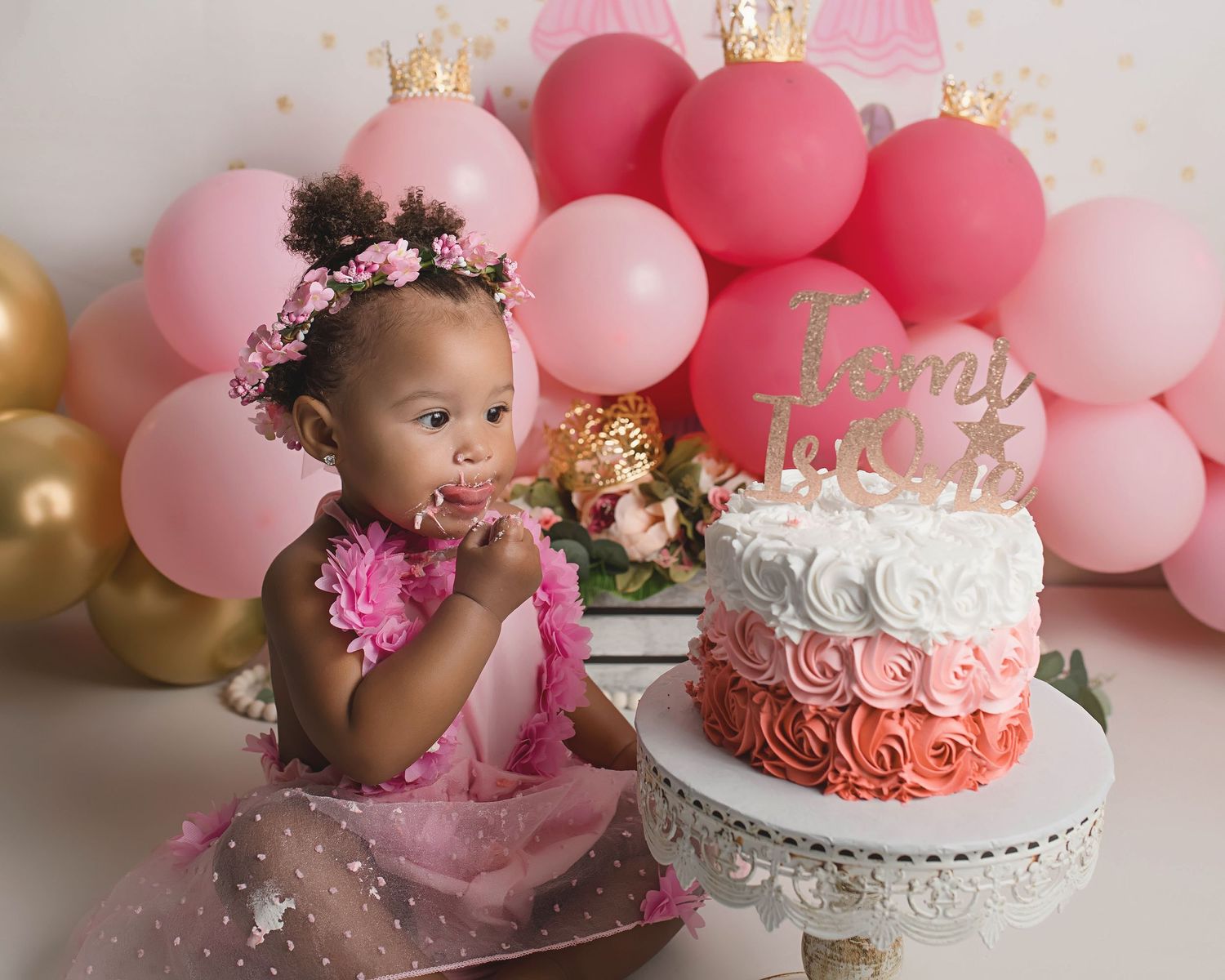 Birthday girl in pink dress enjoying cake smash session with balloon decorations.