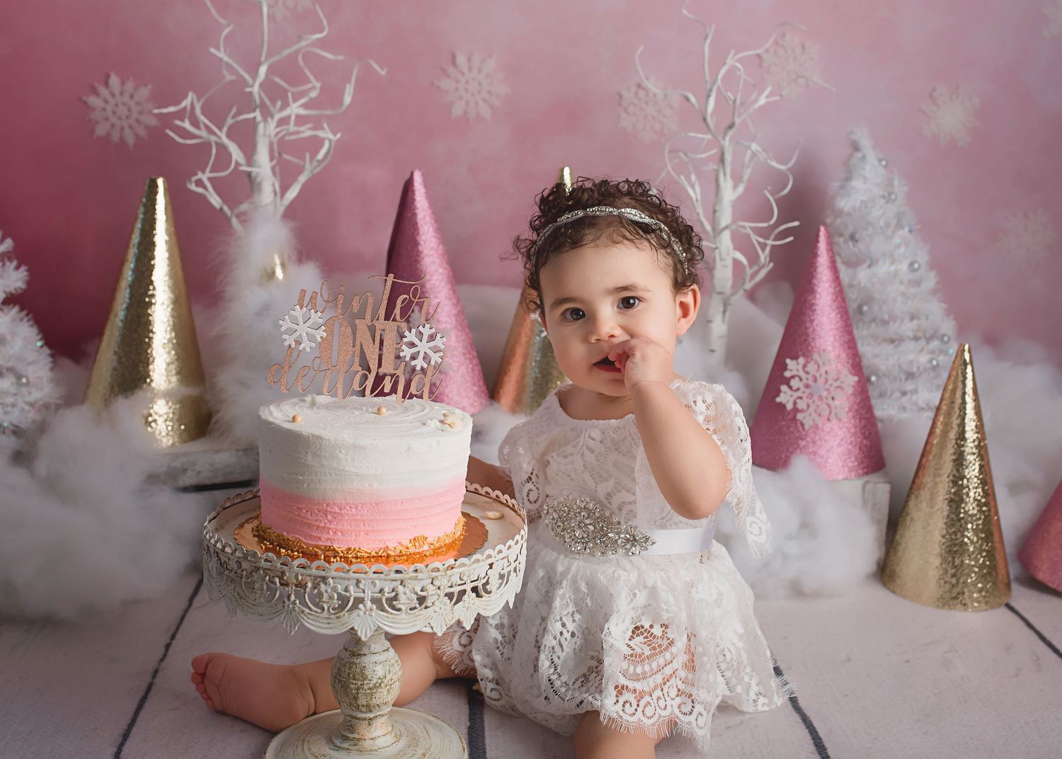 A festive cake smash photo with pink and gold decorative cones and white trees in the background.