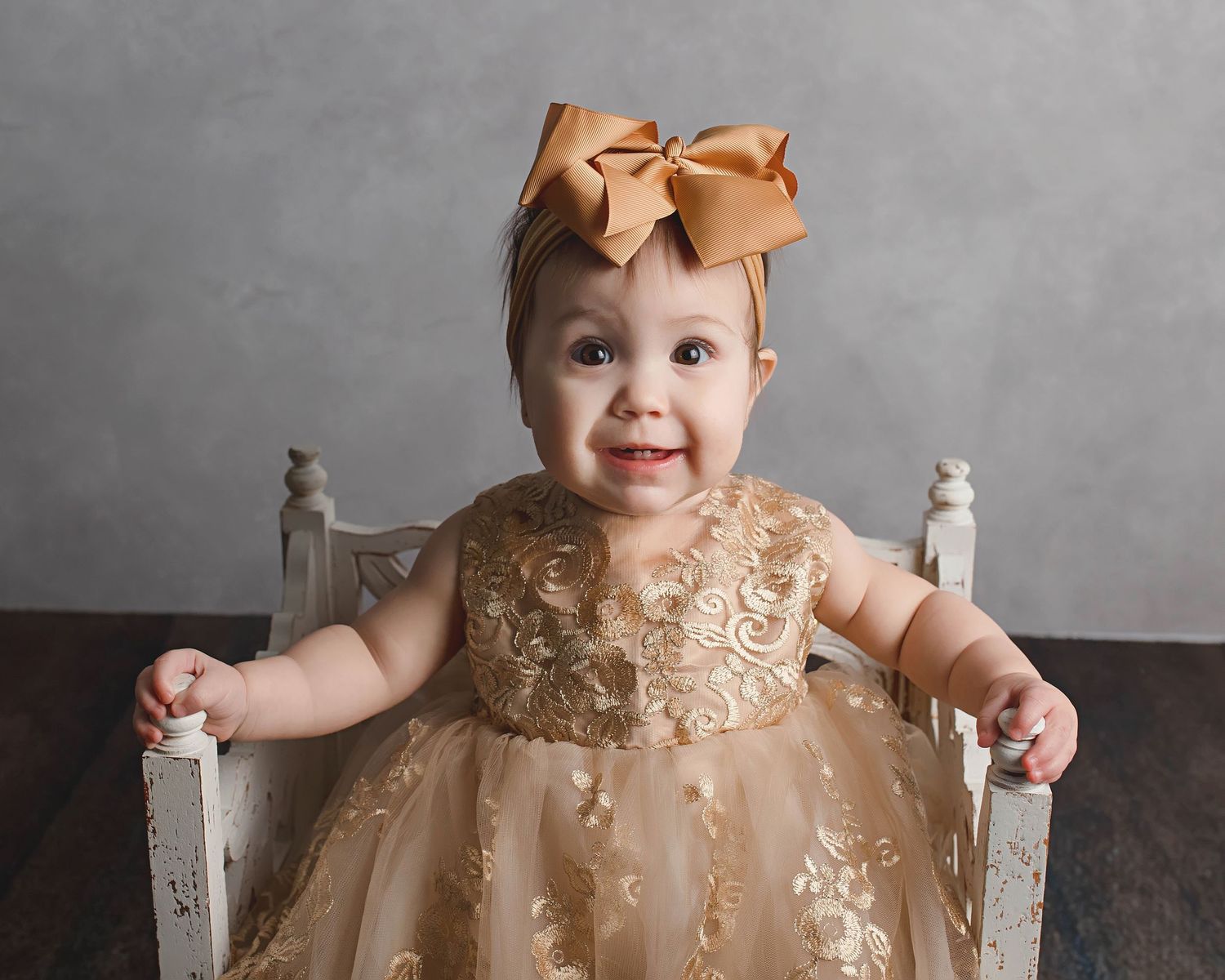 Baby sitting in vintage white chair wearing gold sparkly dress and matching bow.