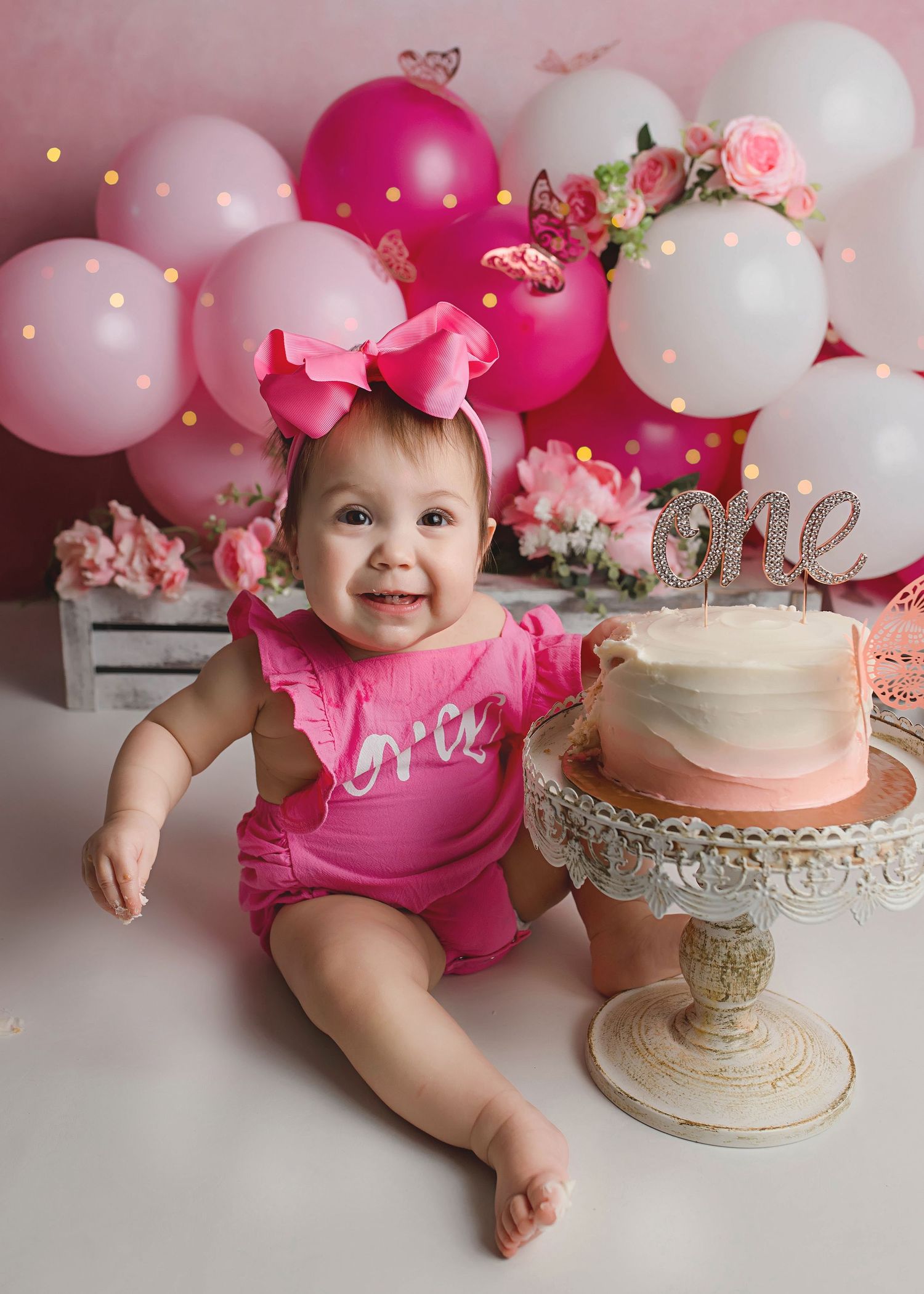 First birthday setup with pink balloons, cake on pedestal, and floral decorations.