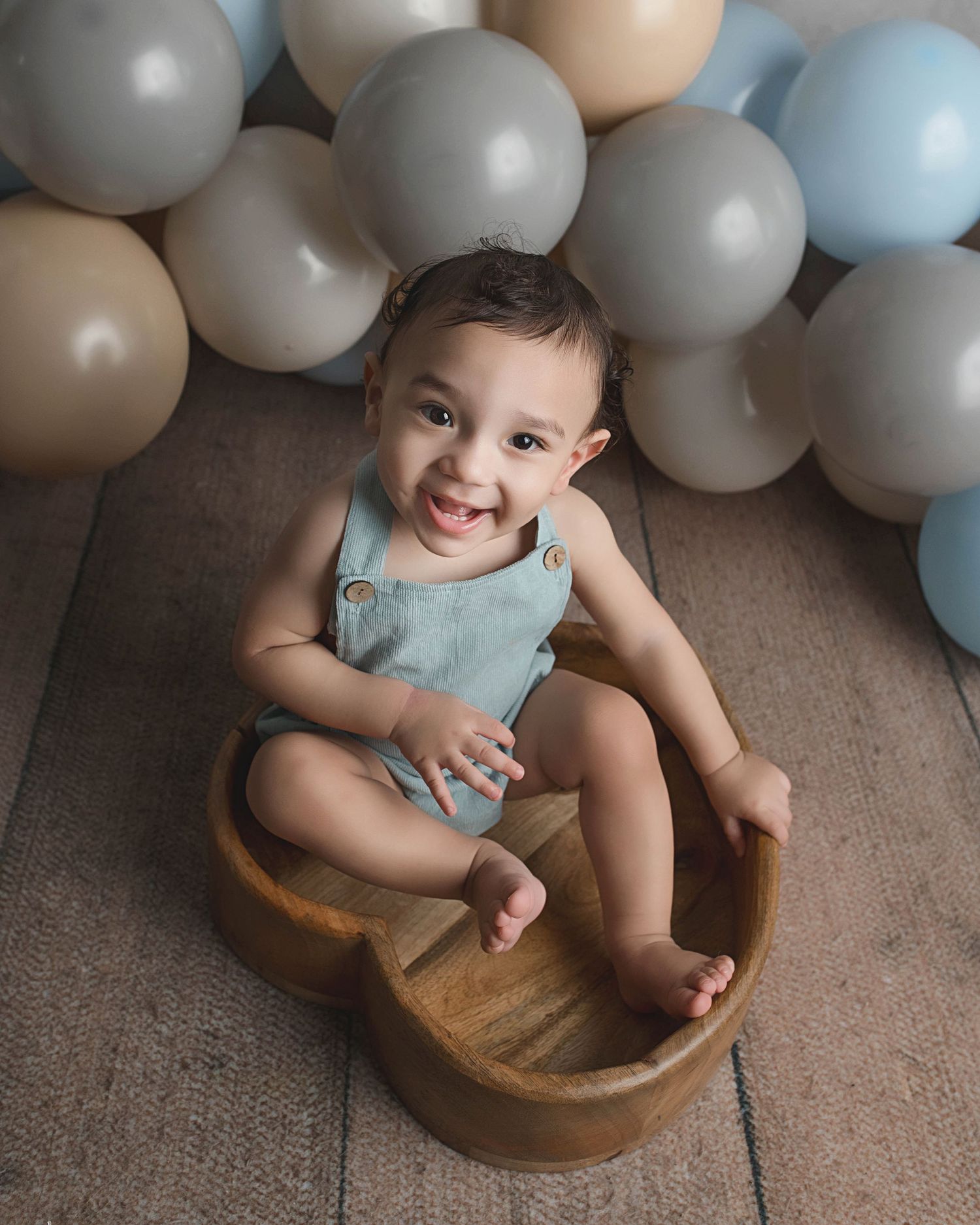 Baby sitting in wooden basin with blue and neutral balloons in background.