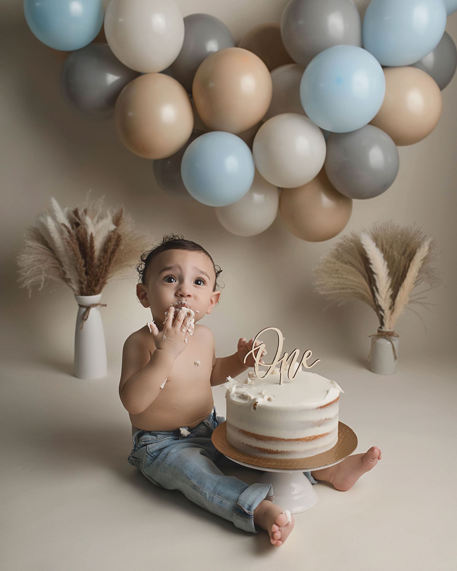 First birthday cake smash photo with blue and neutral balloons, pampas grass decor and white cake.